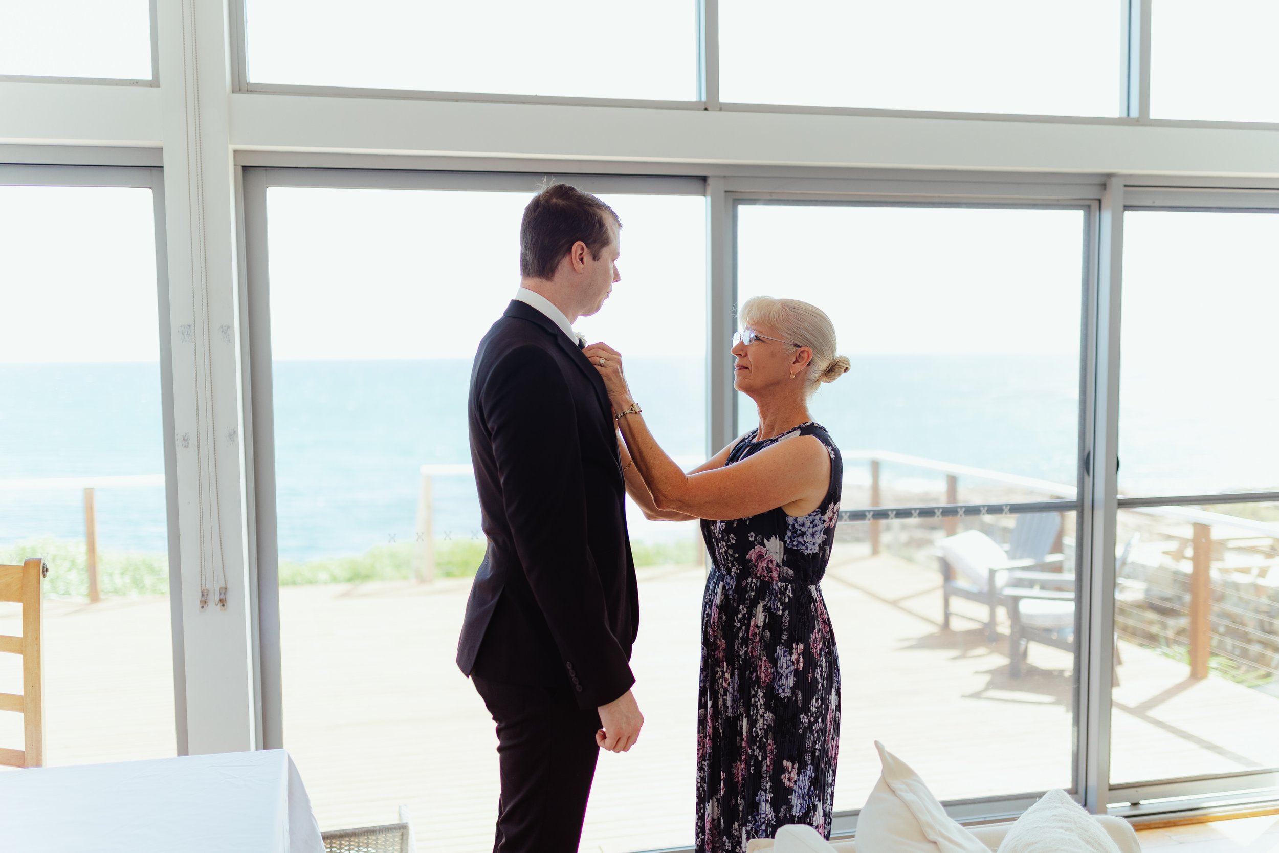 A young man and an elderly woman dance together in a bright room with large windows and a view of the ocean, during daytime.