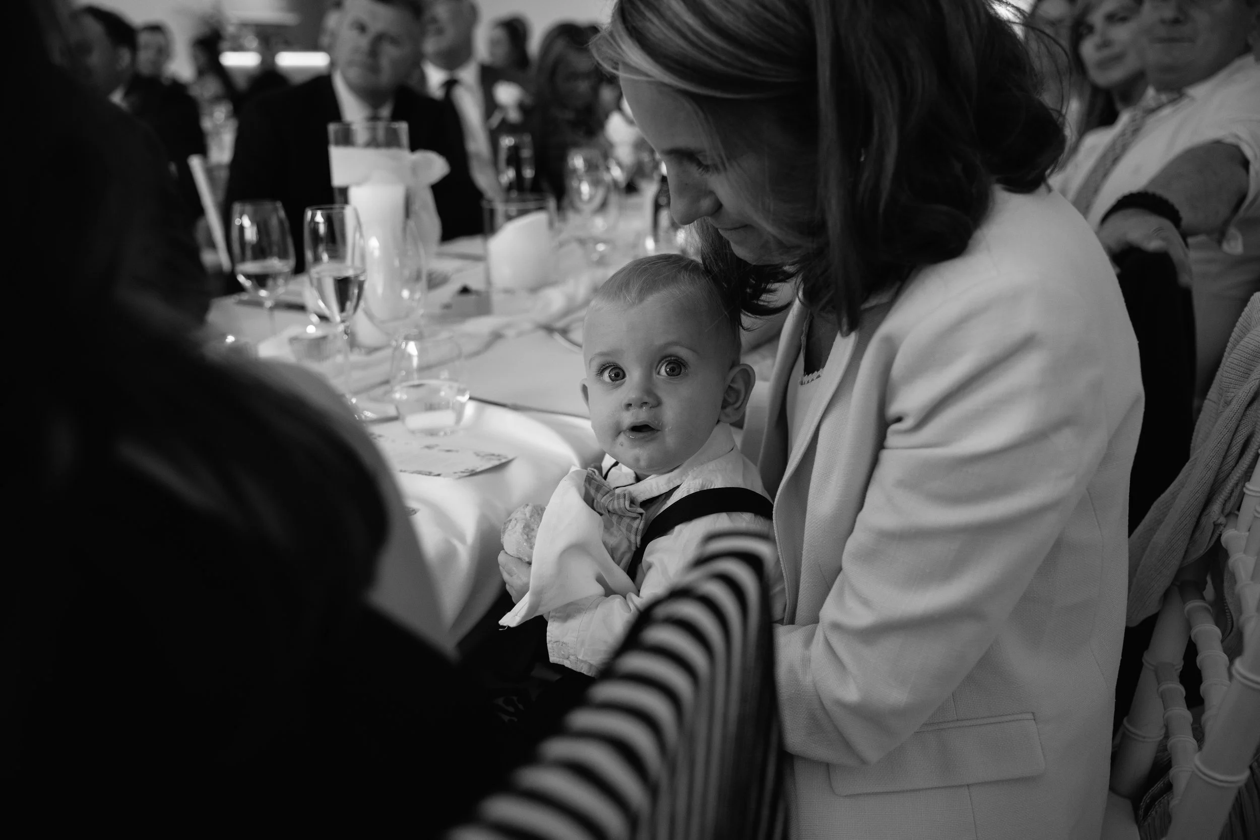 A woman with wavy hair smiling and looking down at a young boy with big eyes, sitting at a table in a restaurant or banquet setting, surrounded by other people in formal attire.