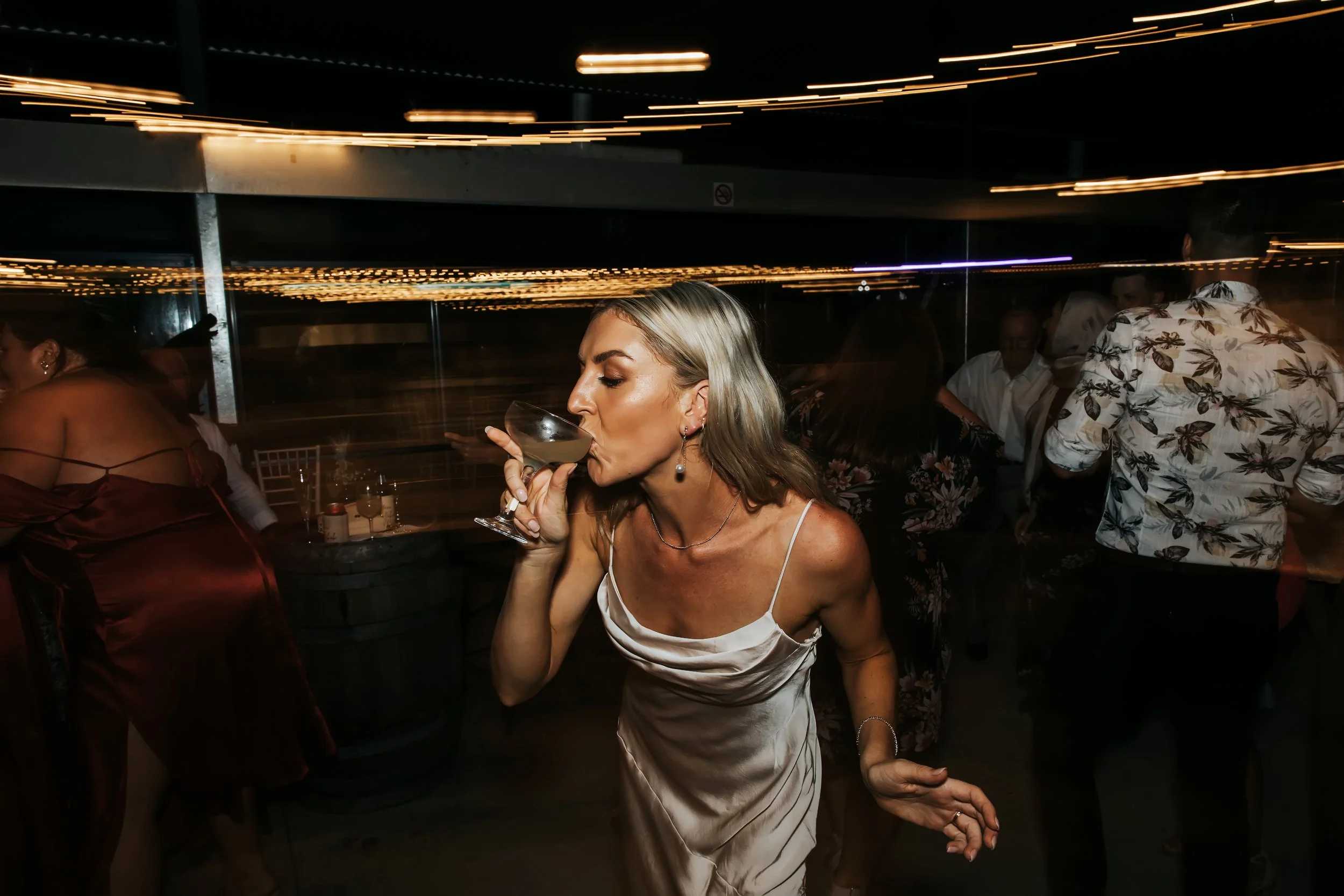 A woman with blonde hair in a white dress drinking from a cocktail at a nighttime social event, with other people in the background and streaks of light from movement.