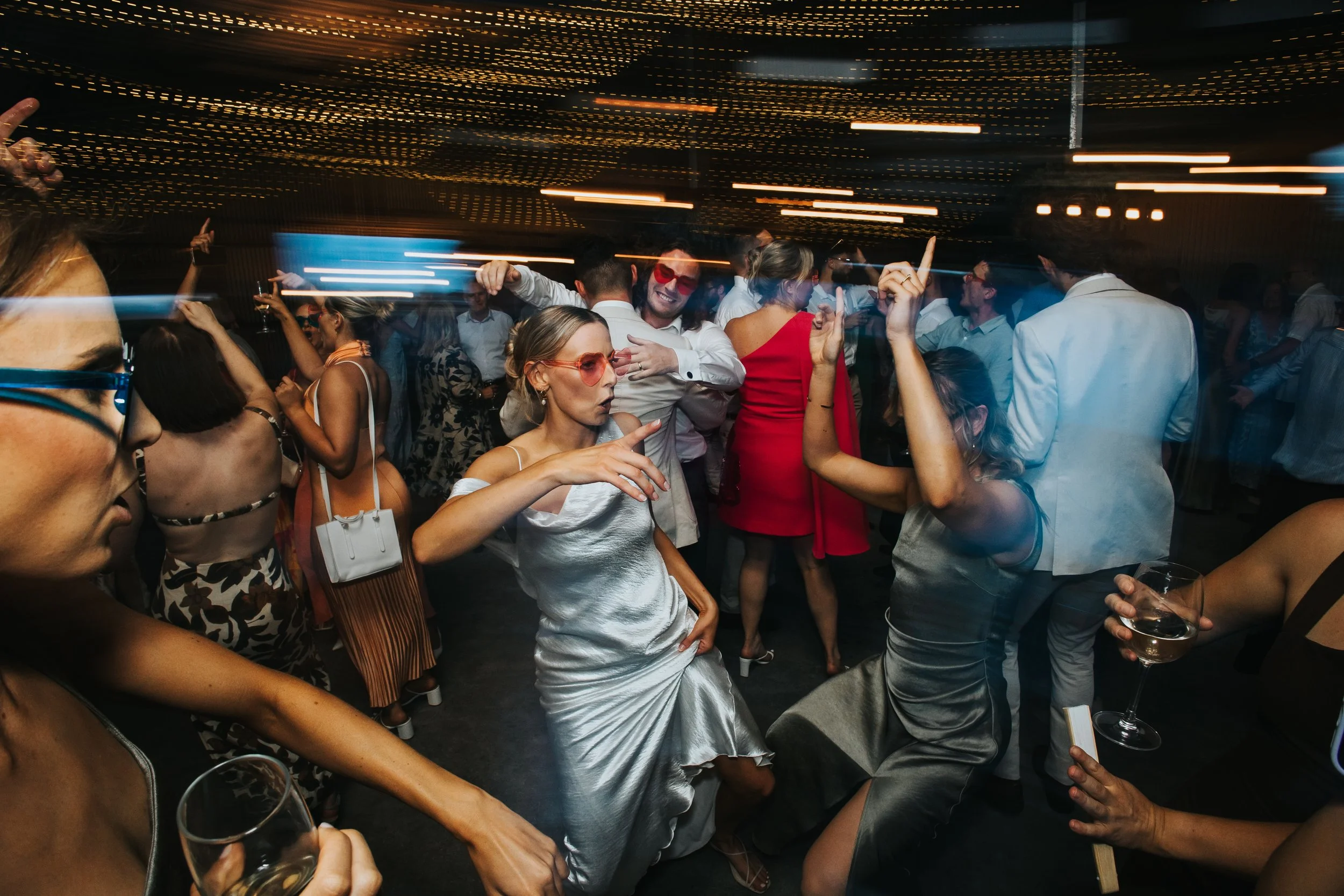 Crowd of people dancing and socializing at a party or nightclub with dim lighting and colorful lights, some holding drinks.