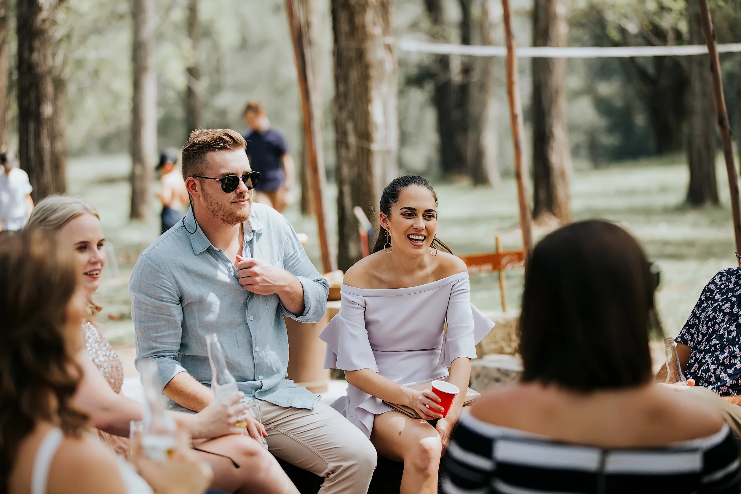 People sitting outdoors at a gathering, some holding drinks, surrounded by trees, enjoying sunlight and conversation.