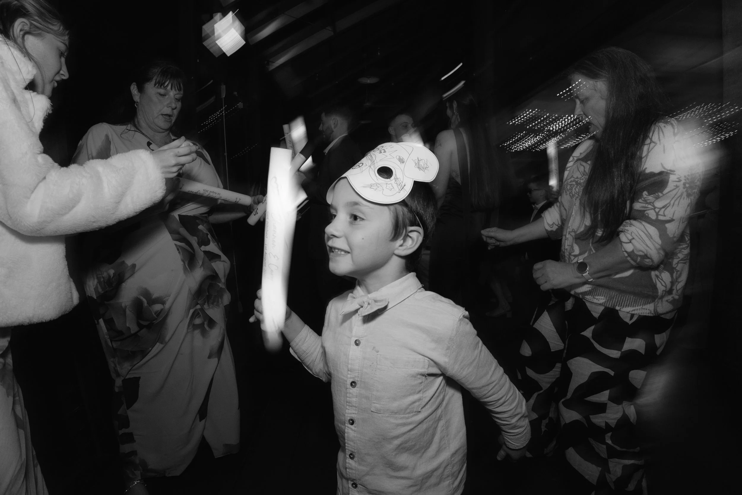 A young boy wearing a paper mask with Halloween-themed drawings, smiling and looking at a woman. The woman, with long dark hair, is facing the boy and appears to be talking to him at a social event. Several people are visible in the background, indic