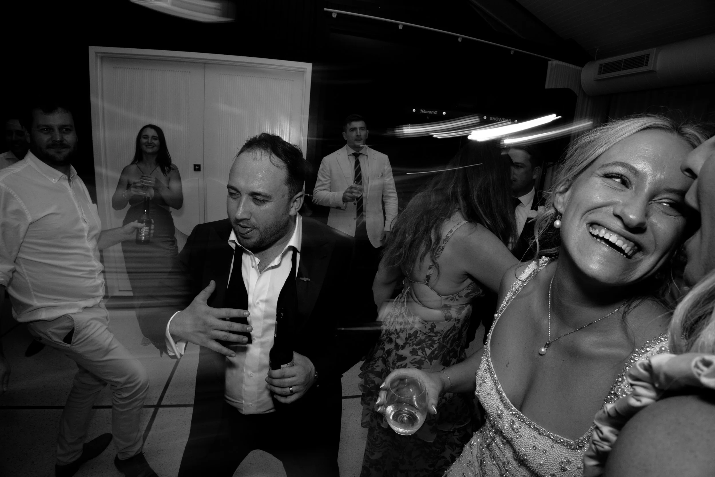 A group of people dancing and socializing at a party, with a woman smiling in the foreground holding a glass of white wine and wearing a sequined dress.