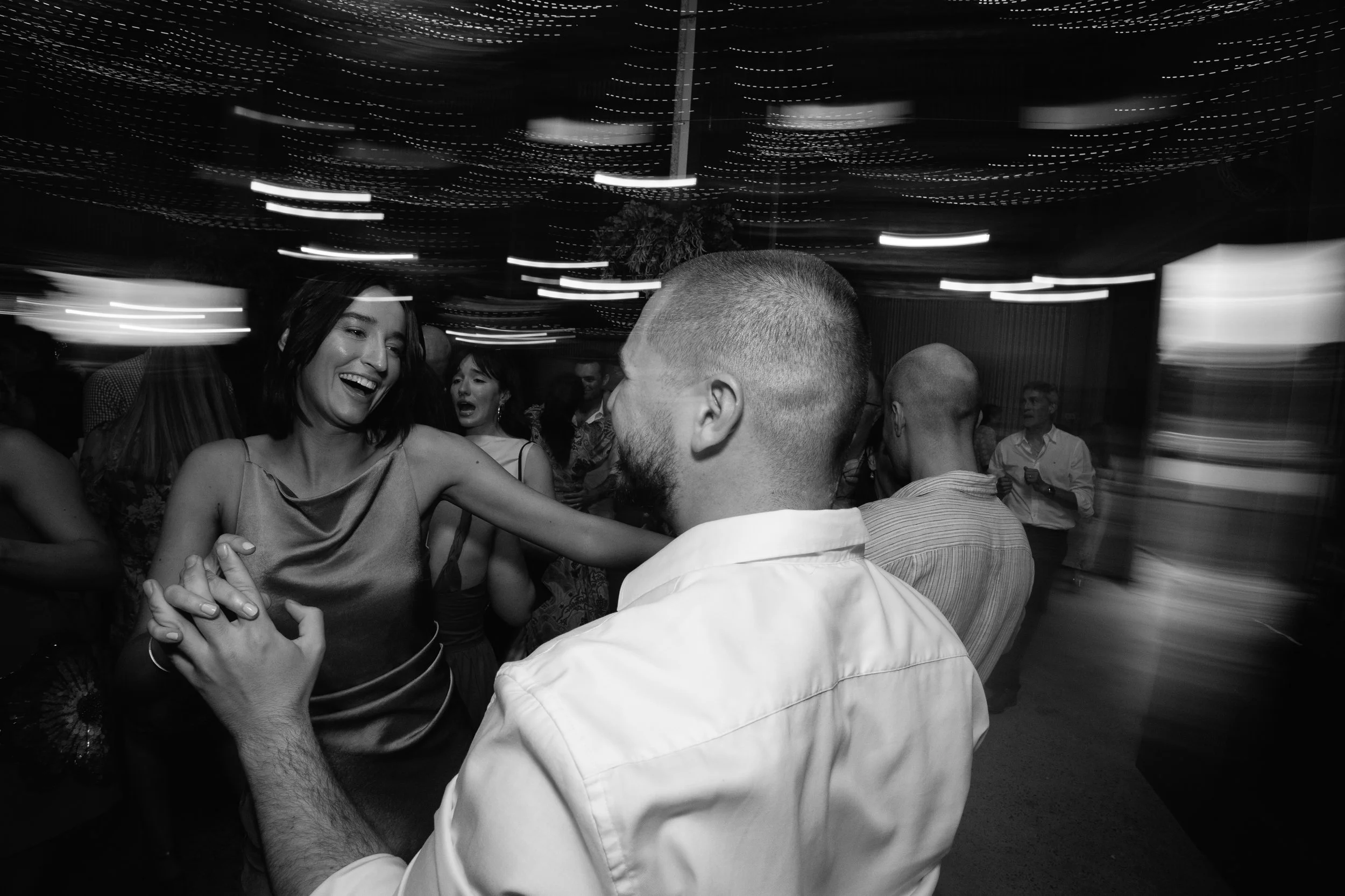 A lively black-and-white photo of people dancing and socializing at a party or celebration, with string lights hanging from the ceiling creating a festive atmosphere.