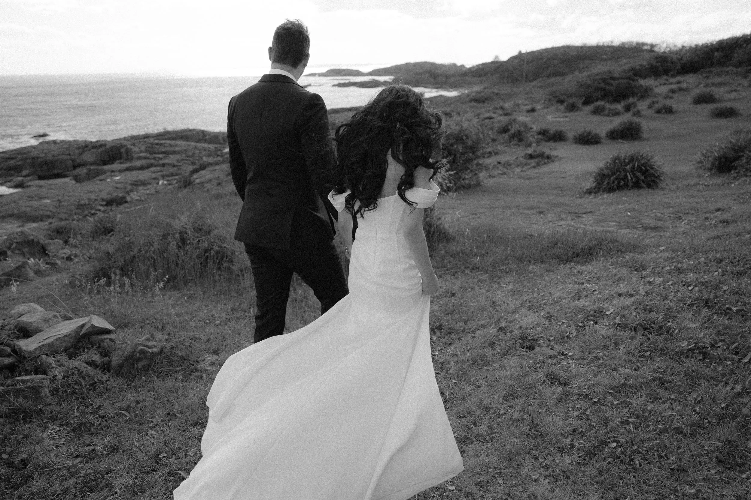 A black and white photo of a man in a suit and a woman in a wedding dress walking away along a coastal landscape, with water and rocky terrain in the background.