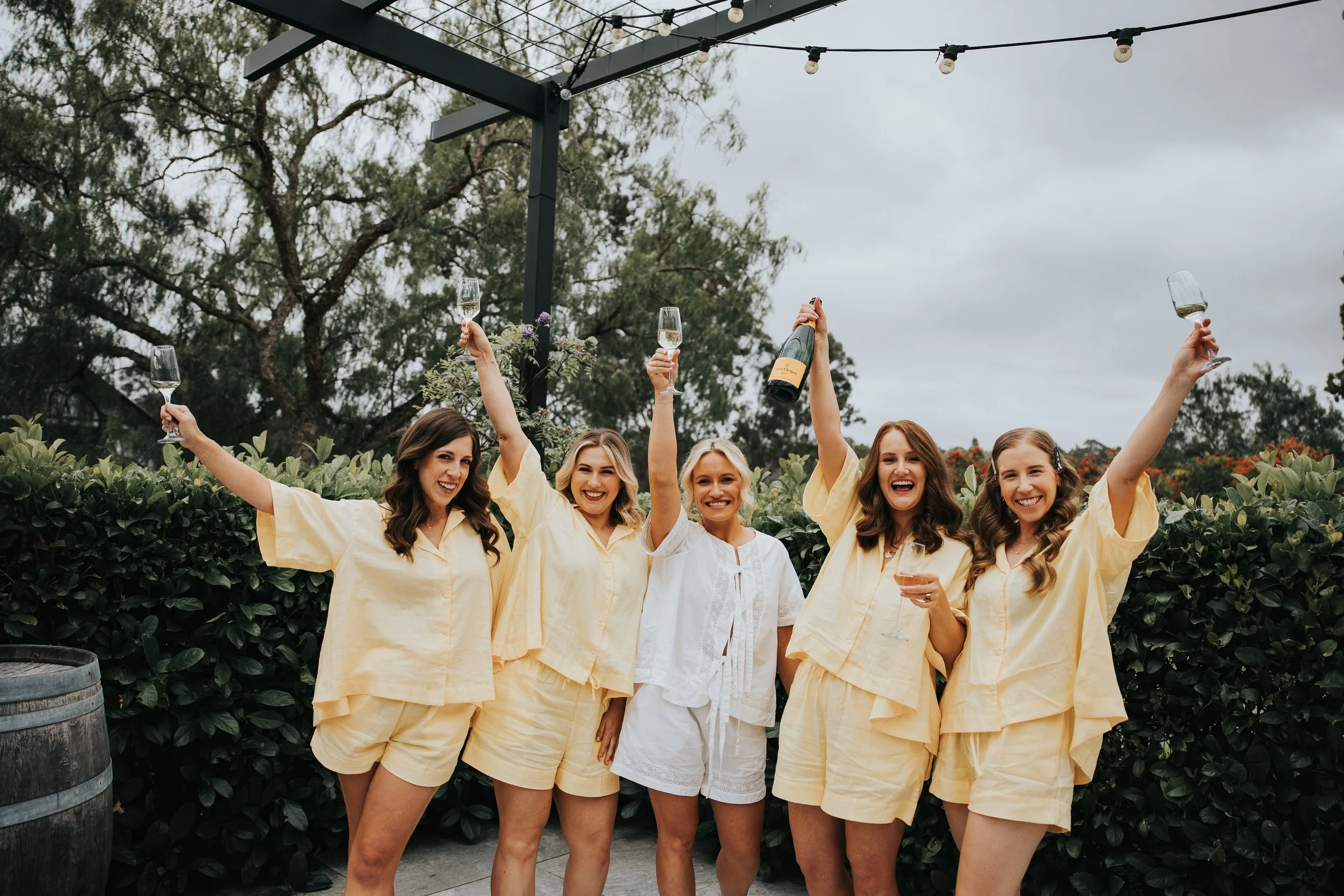 Group of five women dressed in light yellow clothing celebrating outdoors, holding glasses of wine with trees and string lights overhead.