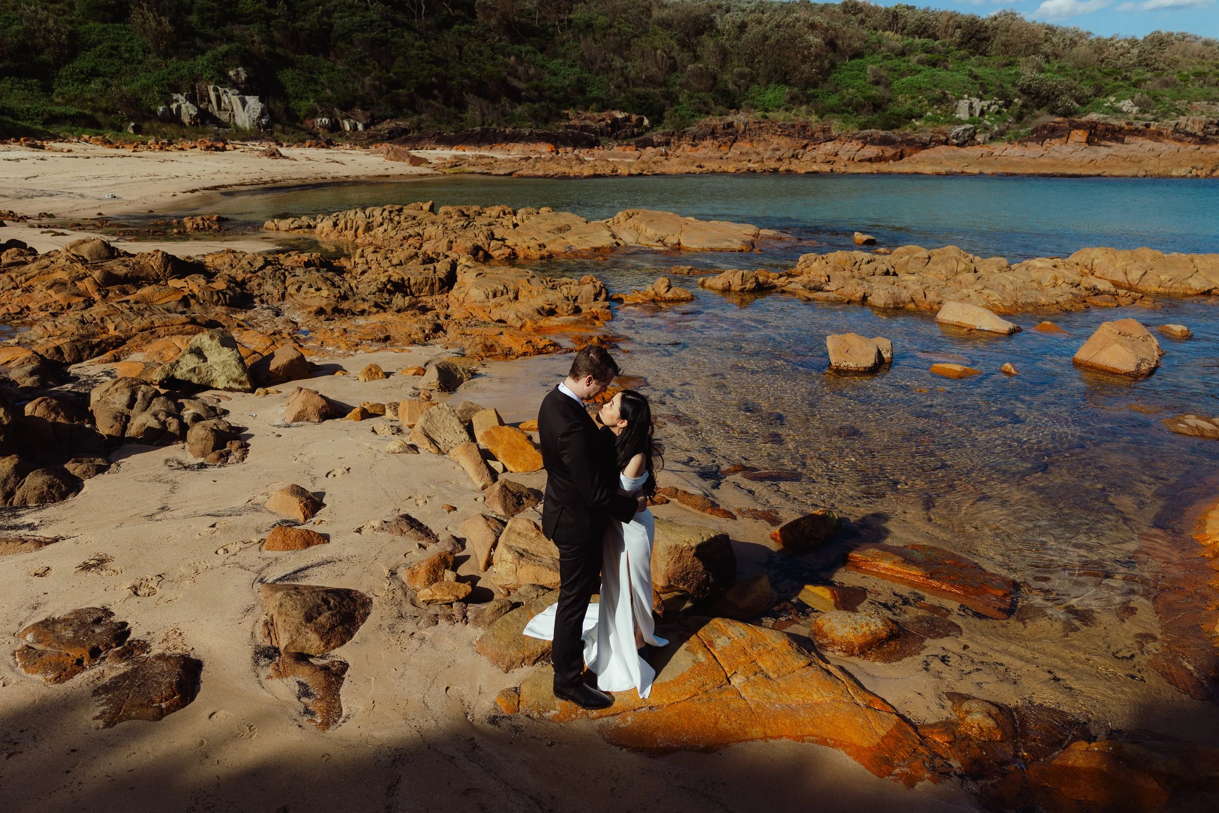 A couple dressed in wedding attire stands on a rocky beach, embracing and gazing into each other's eyes with the ocean and hillside in the background.