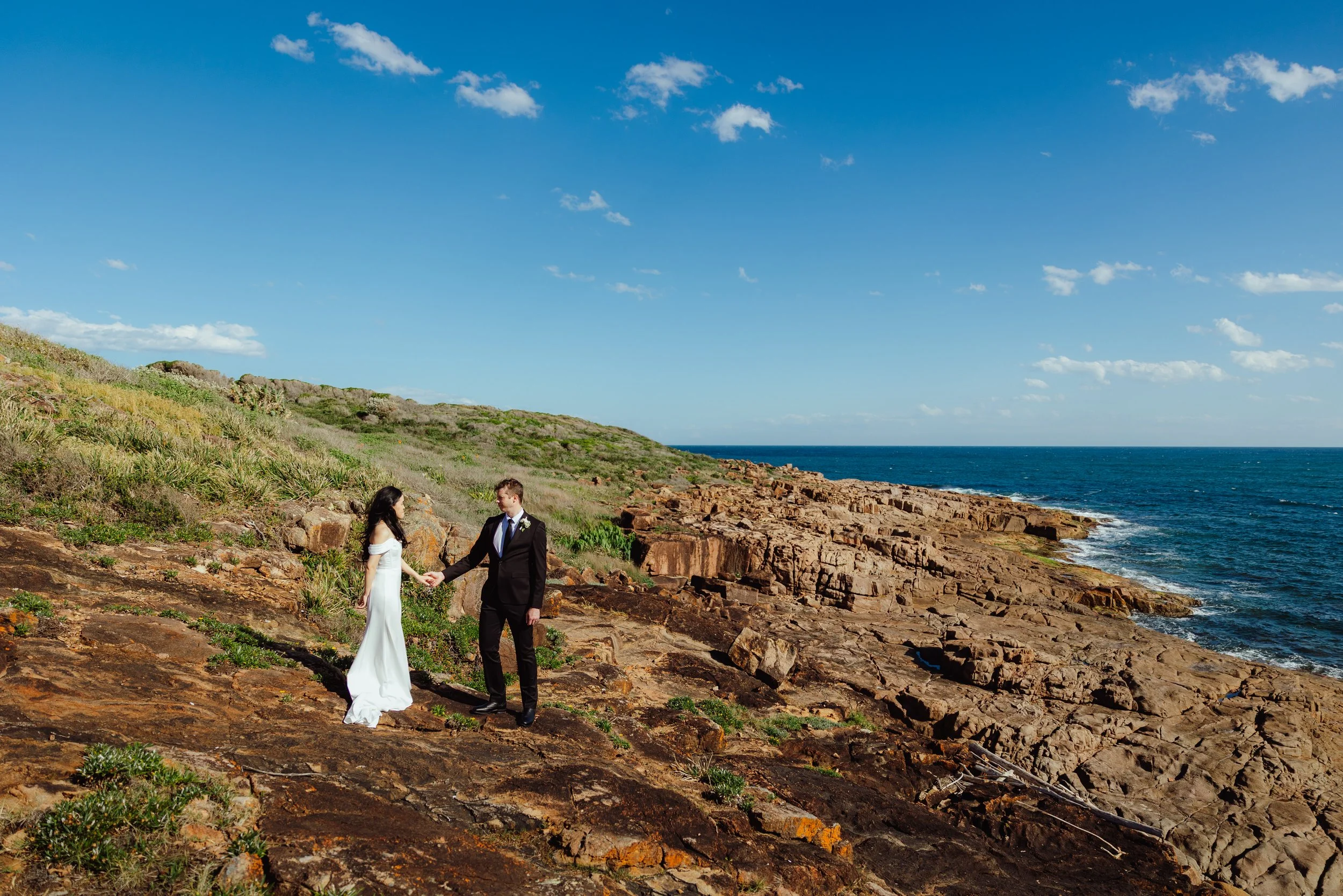 A couple dressed in wedding attire holding hands on a rocky seaside cliff with green bushes and grass, overlooking the ocean and a partly cloudy blue sky.