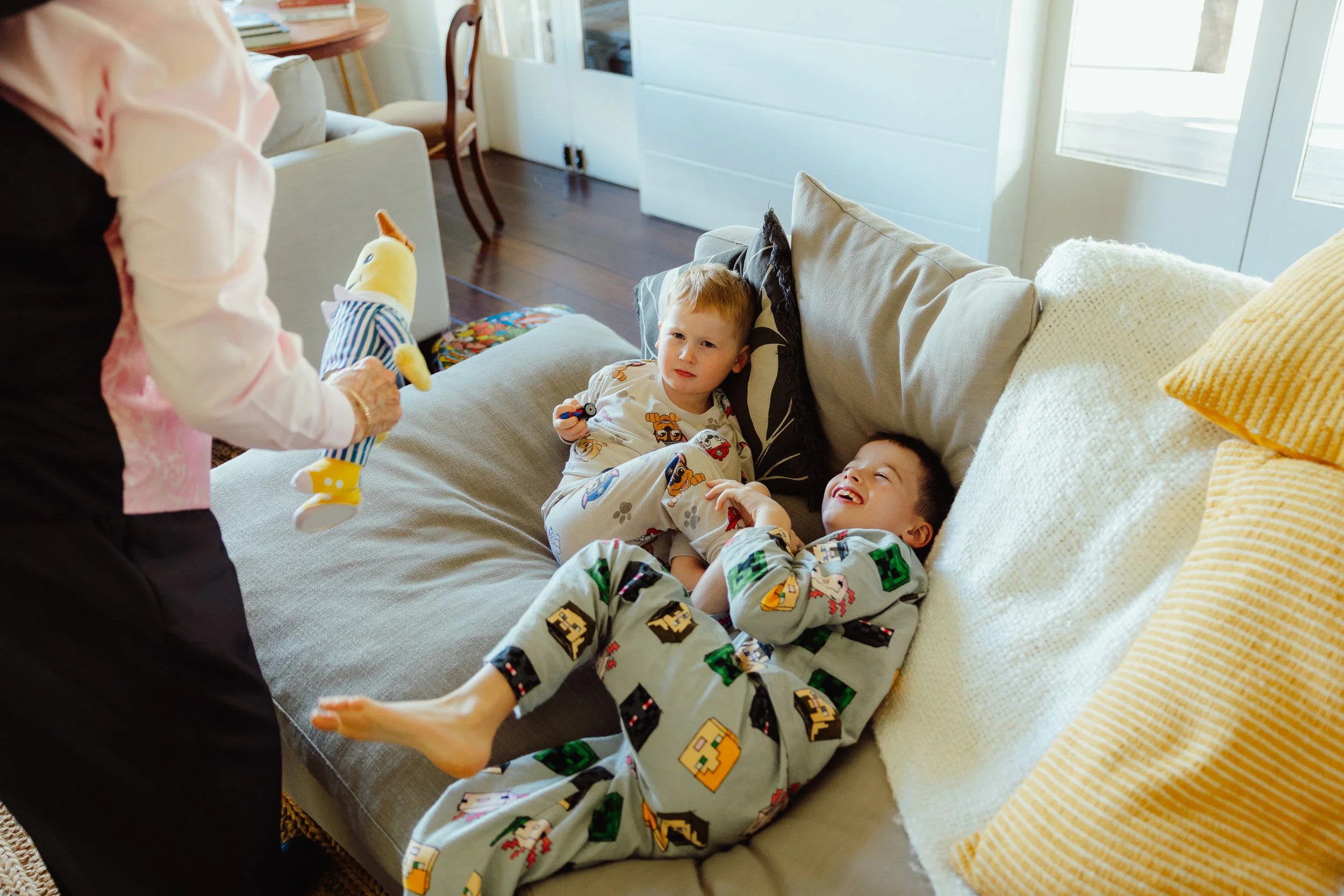 Two young boys in pajamas sitting on a couch, smiling, while a woman holding a stuffed toy shows it to them in a cozy living room with natural light coming through the door.
