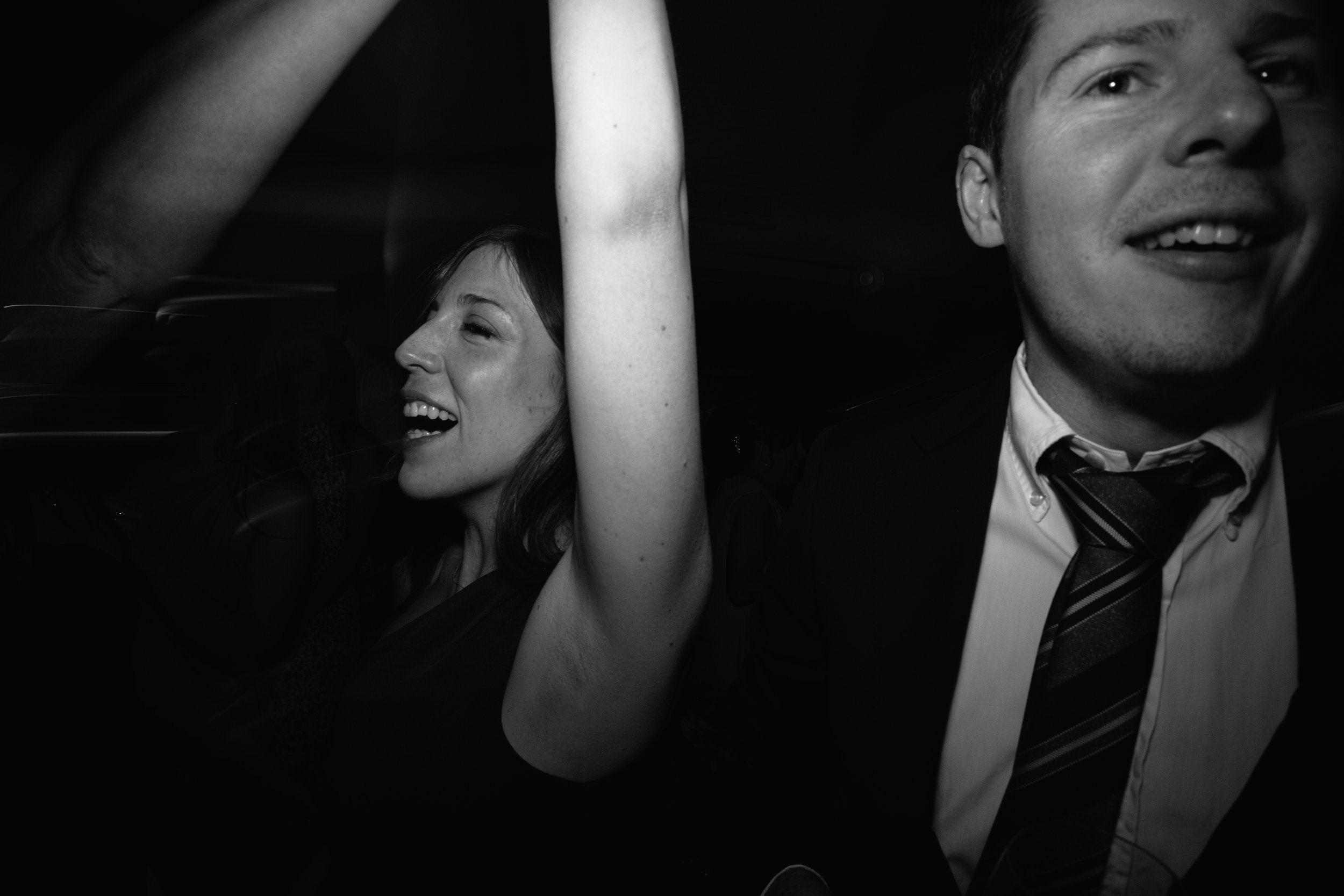 A black and white photo of a man and a woman sitting in a car. The man is on the right, wearing a suit with a striped tie, smiling and looking towards the camera. The woman is on the left, smiling with her eyes closed and arm raised above her head.