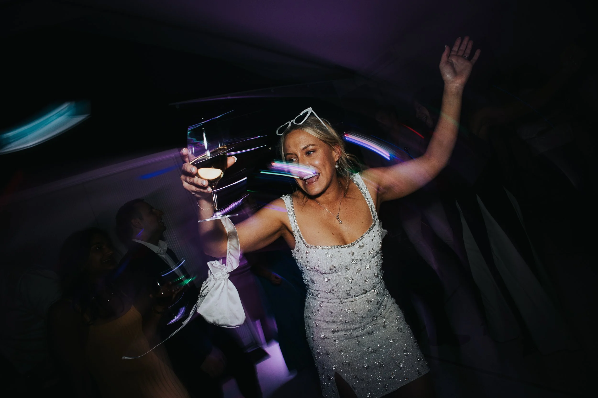 A woman in a white beaded dress dancing at a party, holding a glass of wine, with glasses on her head, surrounded by colorful lights and other people.