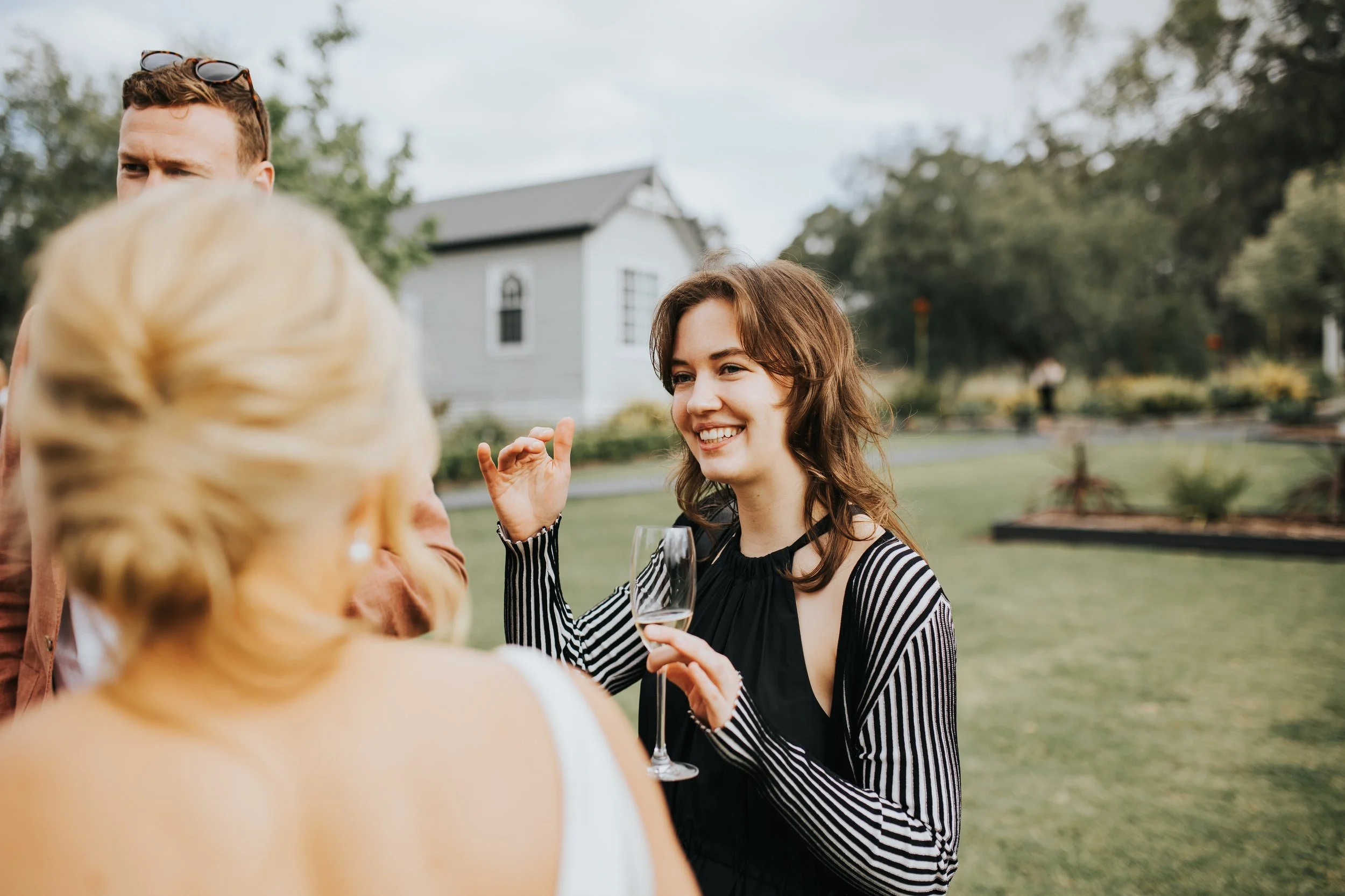 People socializing outdoors during daytime, with a woman smiling holding a glass of wine, and others engaged in conversation, in a garden-like setting with trees and a house in the background.