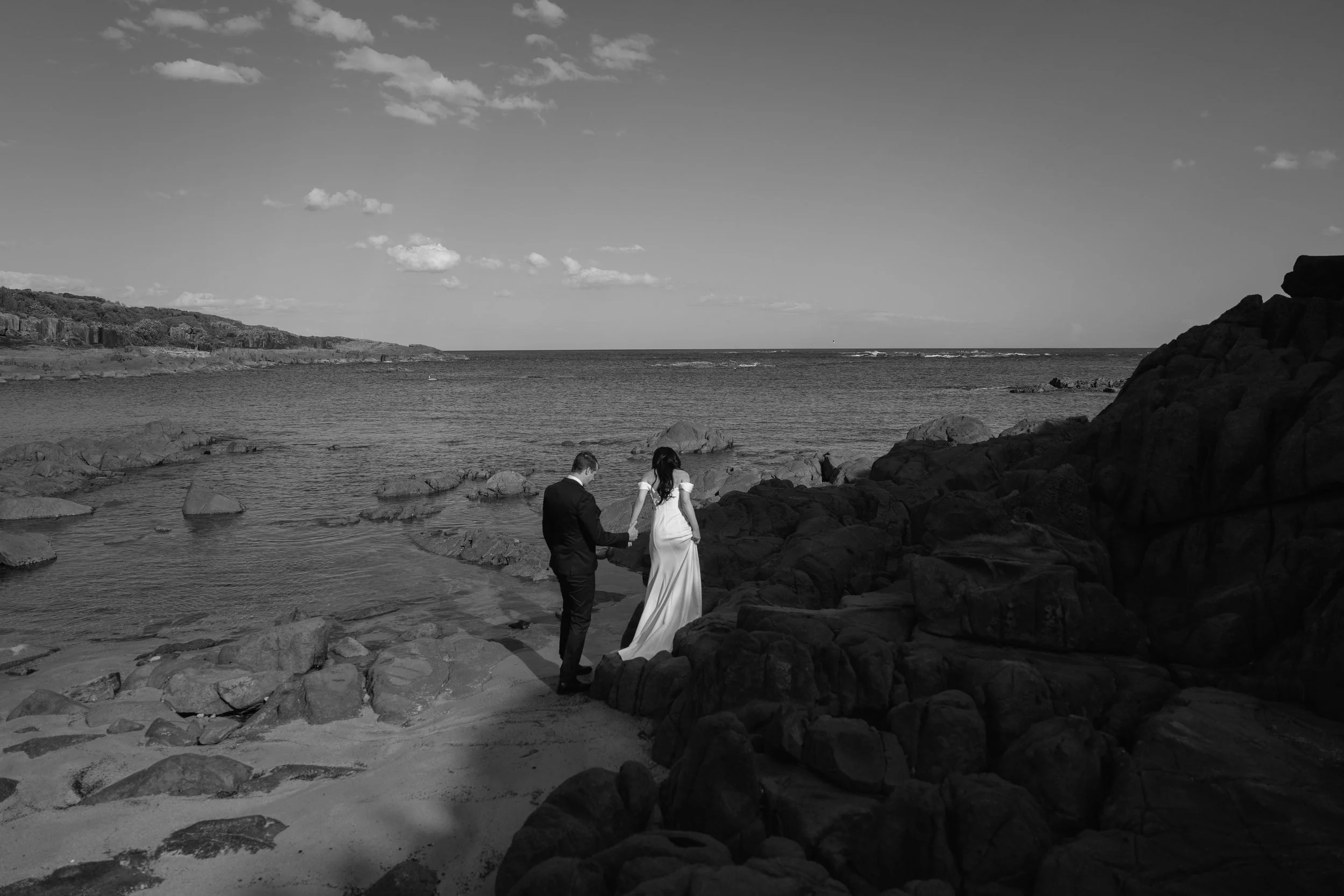 A black and white photo of a couple in wedding attire holding hands at a rocky beach with water and cloudy sky in the background.