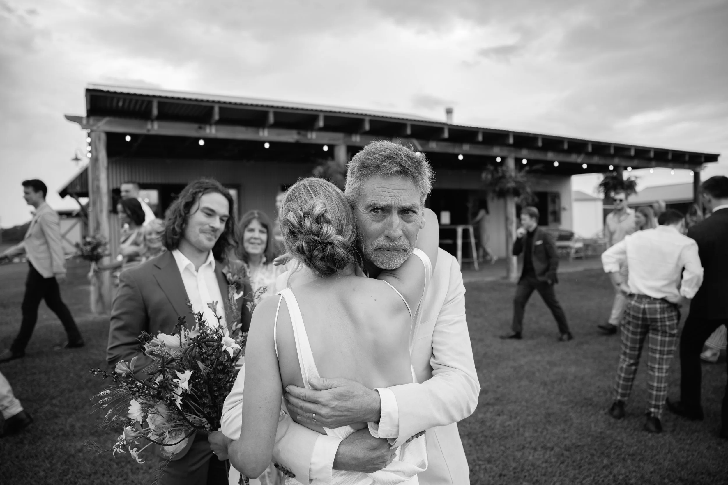 A black-and-white photo of a wedding reception outdoors, featuring a couple hugging. The bride, holding a bouquet of flowers, has her face against the groom’s chest, while the groom looks directly at the camera with a serious expression. People are m