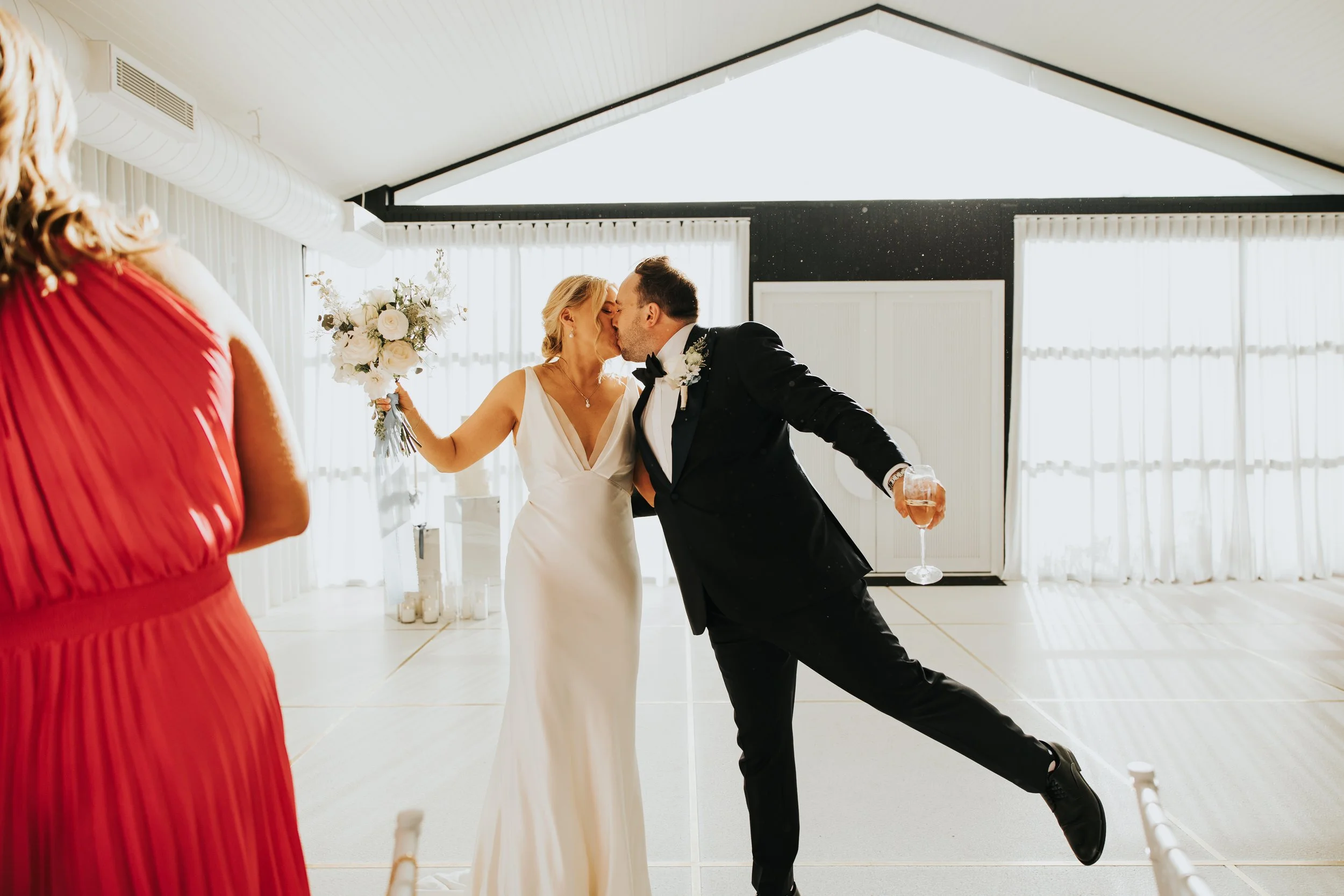 A bride and groom sharing a kiss at their wedding reception, with a woman in a red dress holding a bouquet visible in the foreground.