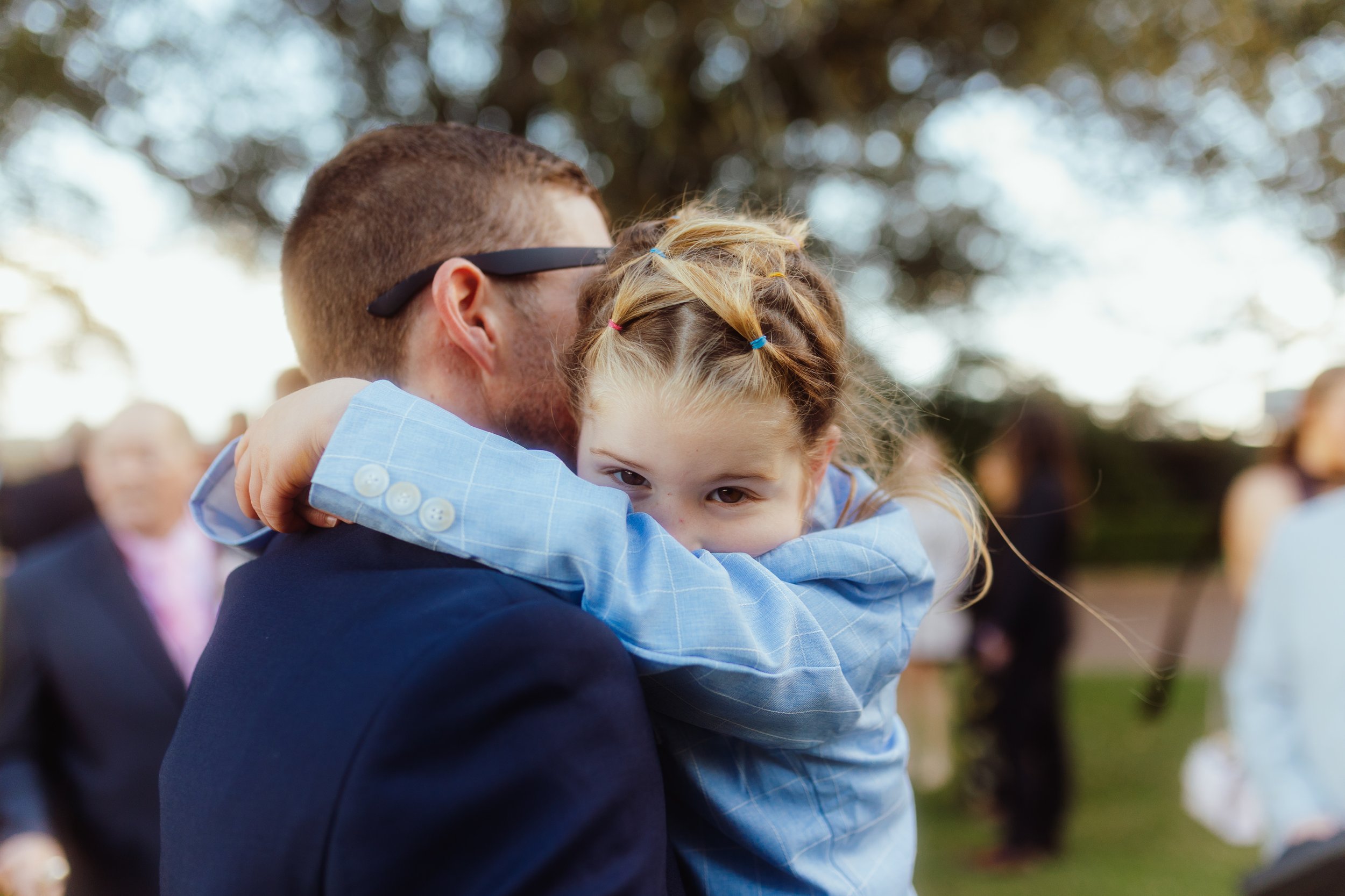 A man with sunglasses embraces a young girl with braided hair, both wearing formal attire outdoors during daytime.