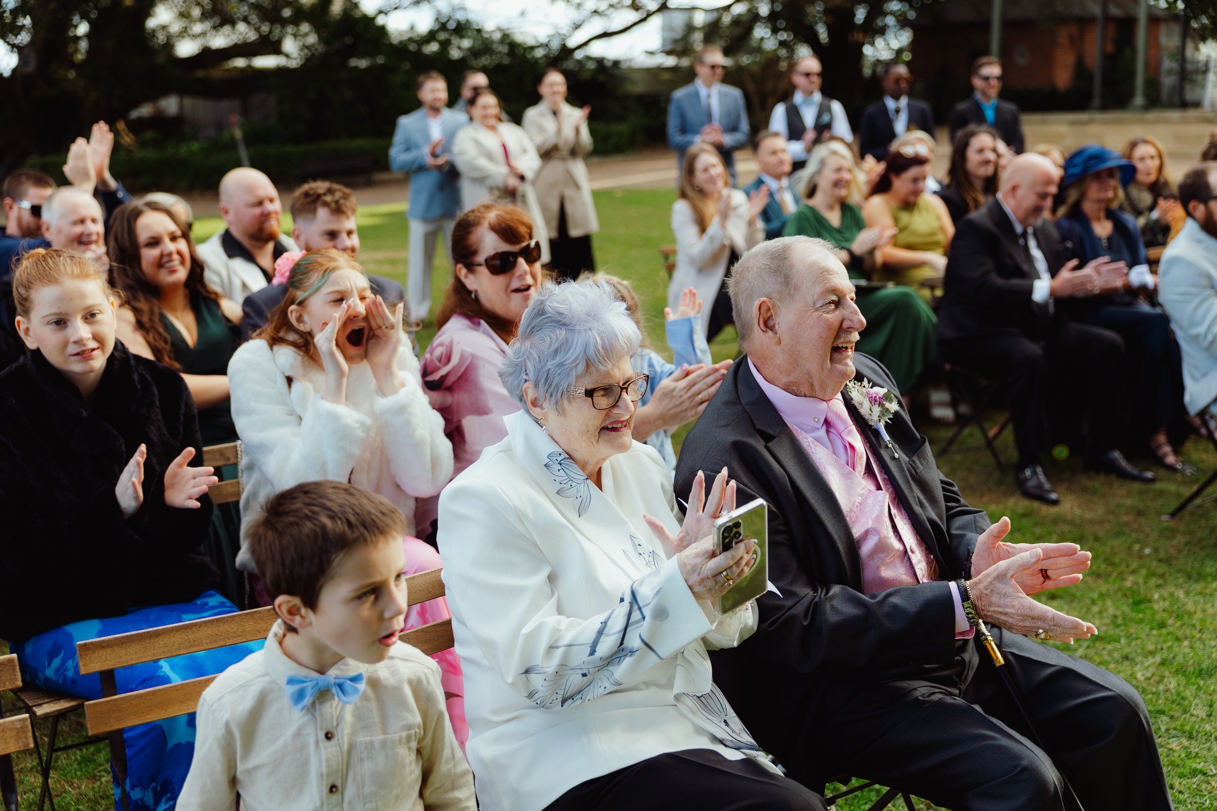 A group of people attending an outdoor event, sitting and standing on grass, engaging with a performance or speech, with trees and a brick building in the background.