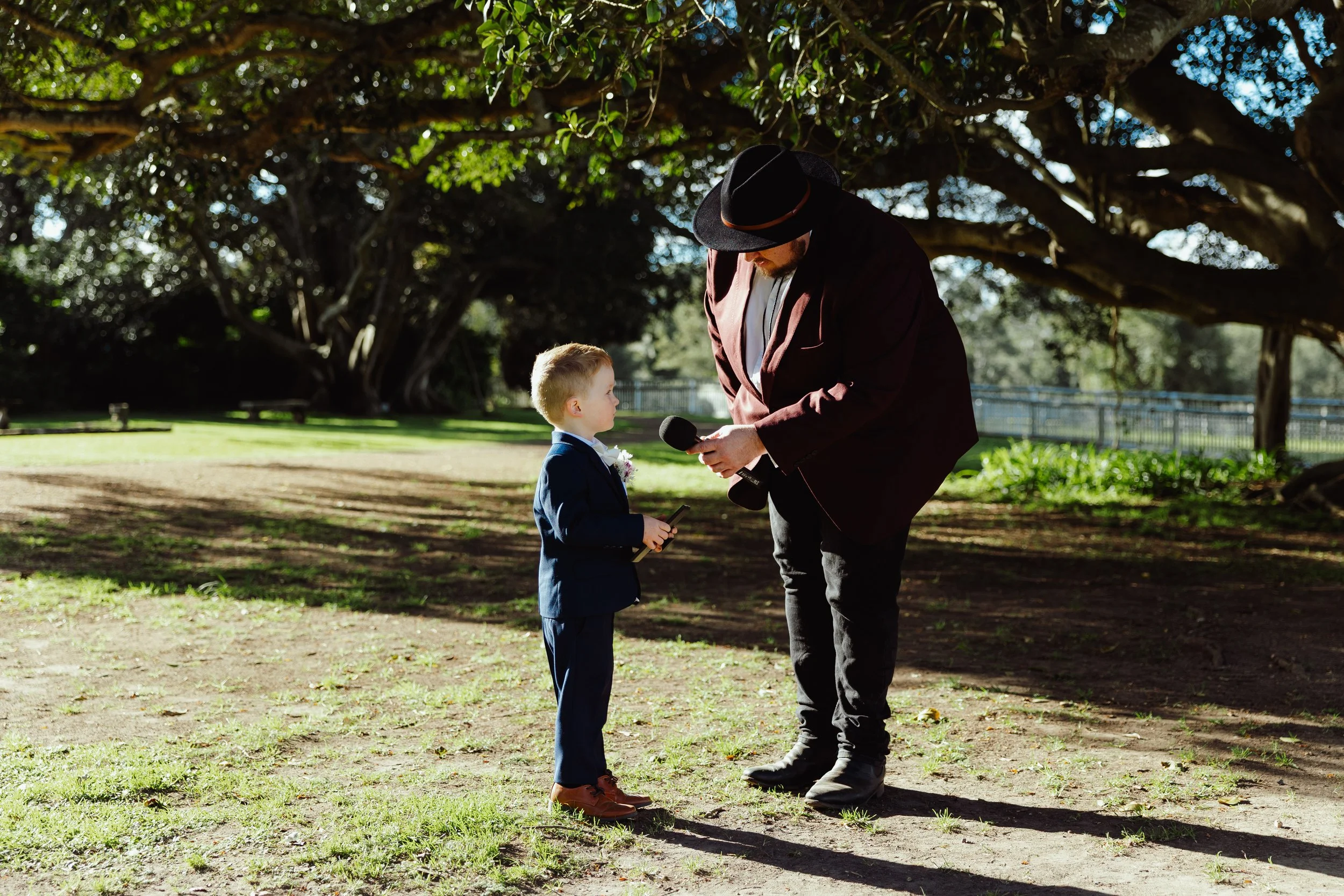 Man dressed in a suit and cowboy hat is holding a microphone and interviewing a young boy wearing a suit and holding a tablet. They are outdoors under large trees with sunlight filtering through the leaves.