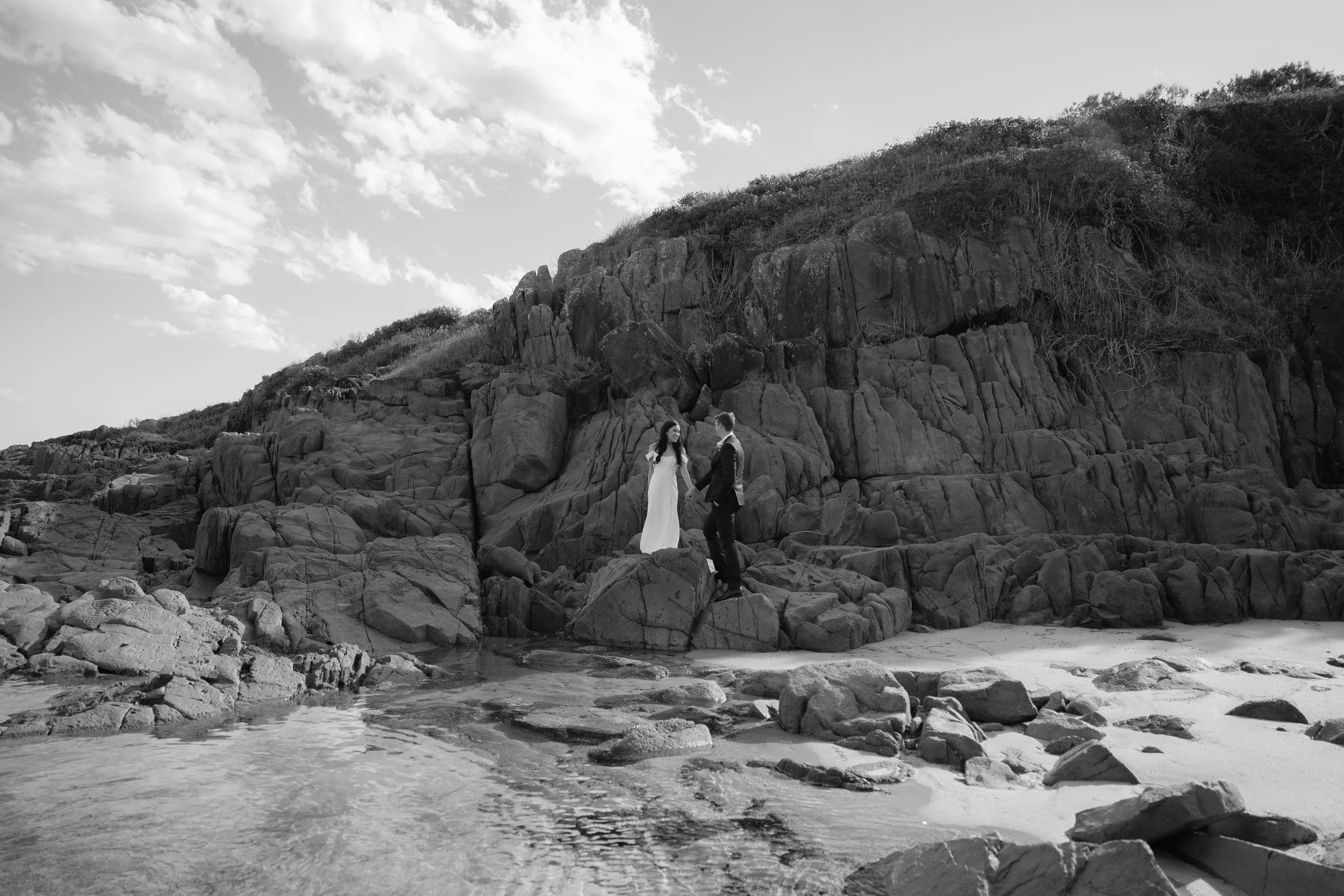 A black-and-white photo of a couple in wedding attire standing on rocks near the water at a beach with large rocks and a hillside in the background.