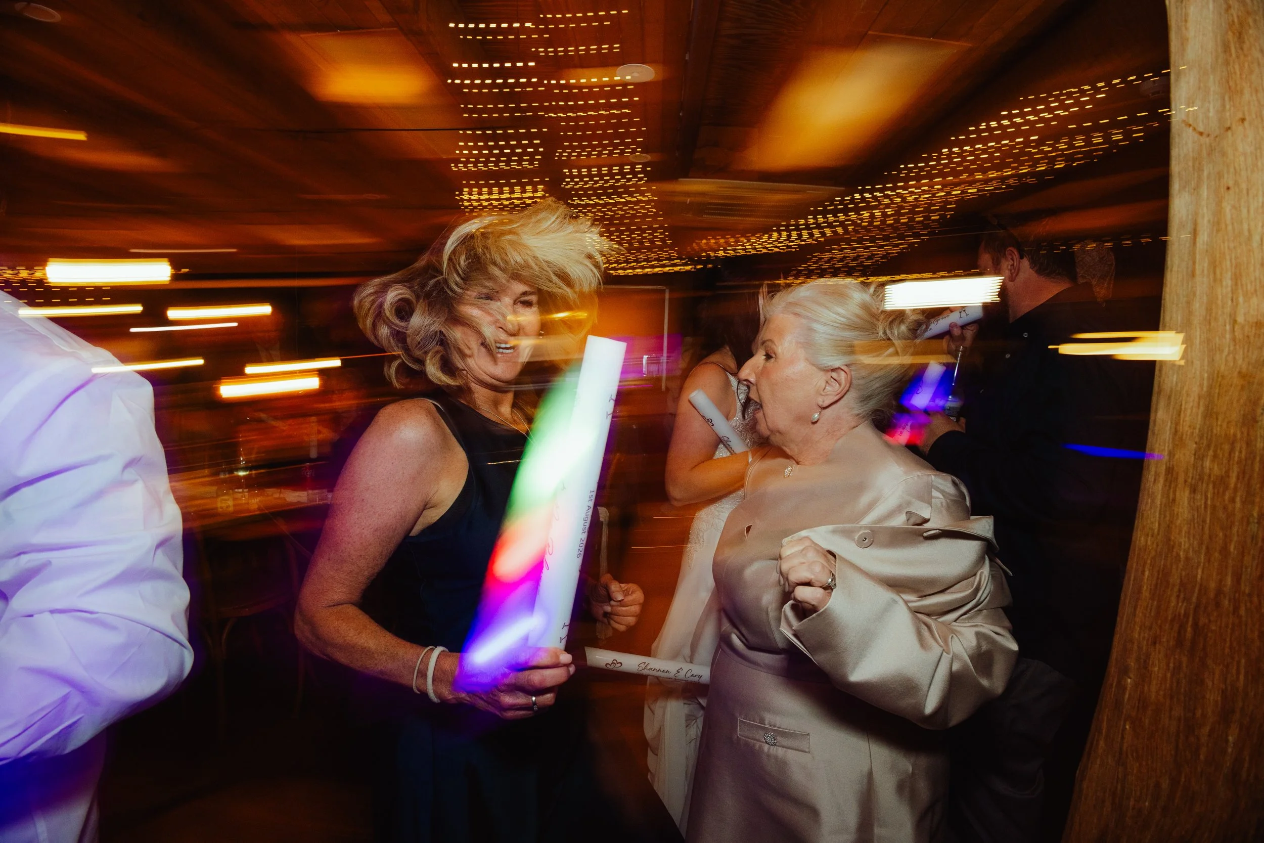 Two women are smiling and sharing a moment at a celebration or party, with string lights in the background creating a lively atmosphere.