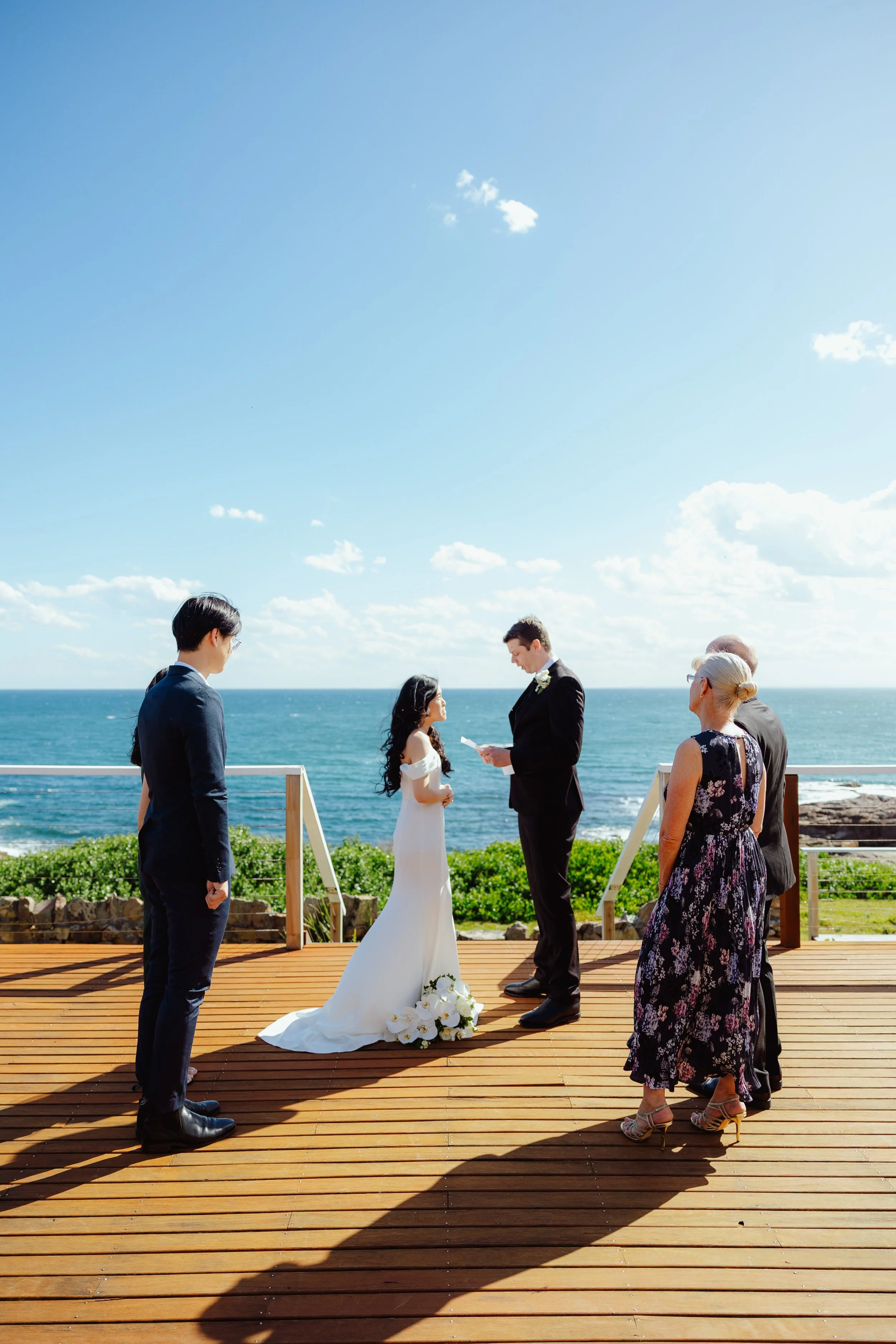 A wedding ceremony taking place outdoors on a wooden deck by the ocean with five people, including a bride and groom exchanging vows, officiant, and guests, under a bright blue sky with clouds.