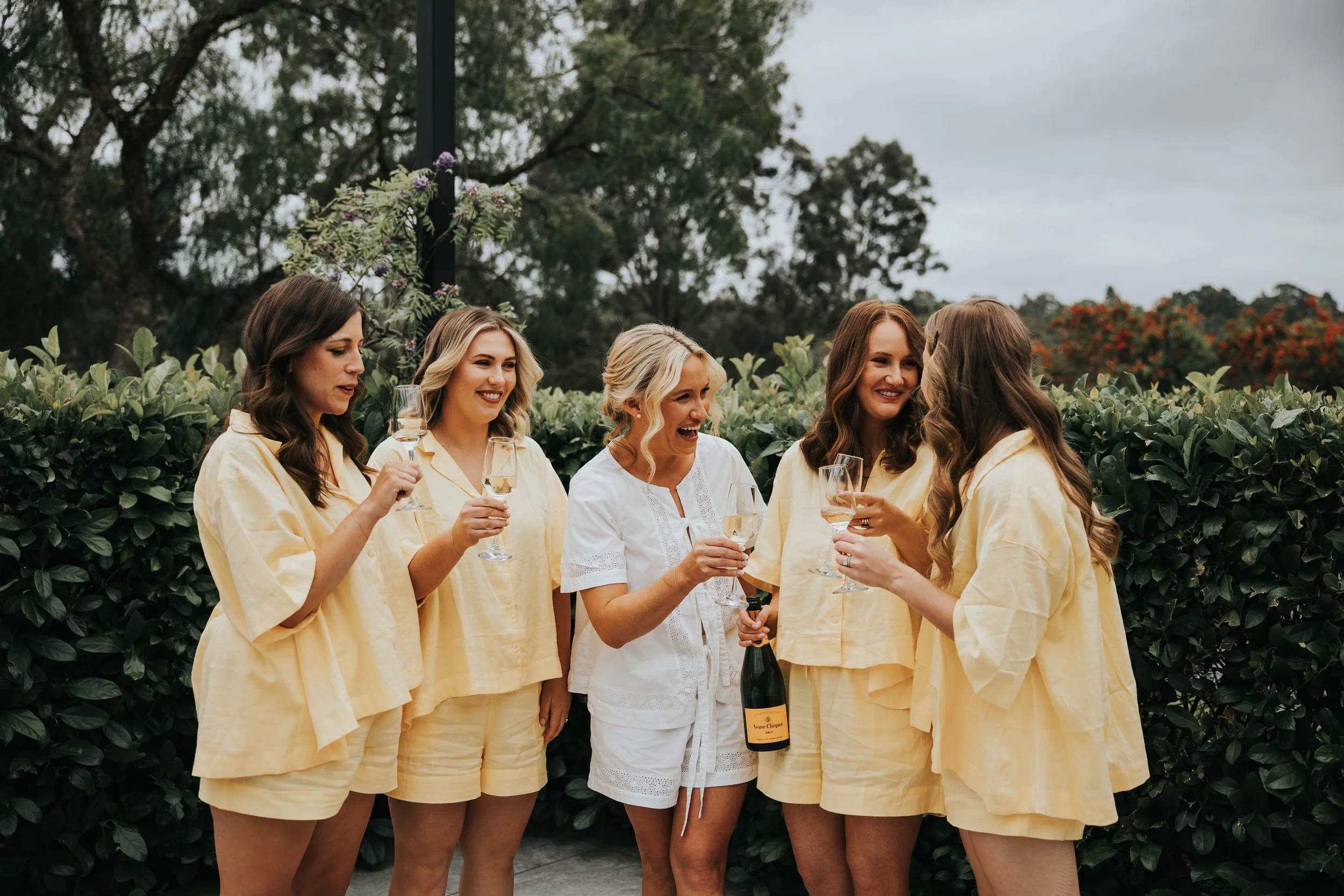 A group of six women wearing matching yellow pajamas, holding glasses of champagne, and smiling while standing outdoors with green shrubbery and trees in the background.