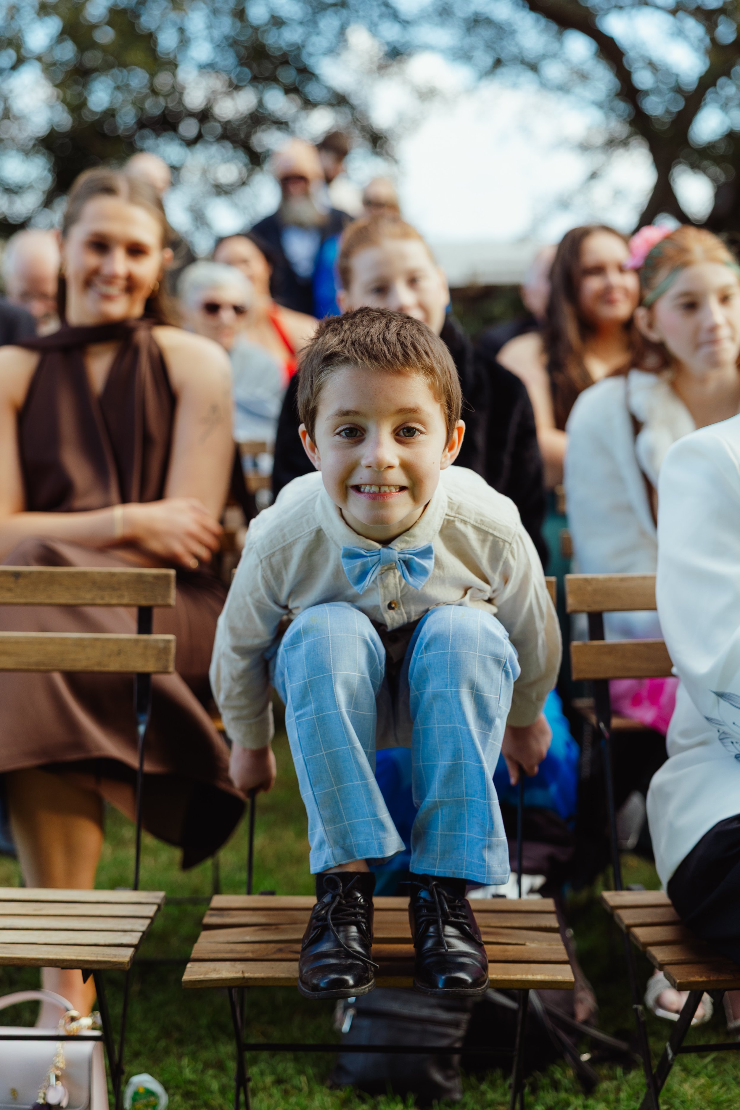 A young boy dressed in a white shirt, blue plaid pants, black boots, and a blue bow tie is sitting on a wooden bench, smiling at the camera. He is at an outdoor event with people sitting behind him, including women and children, under trees with blur
