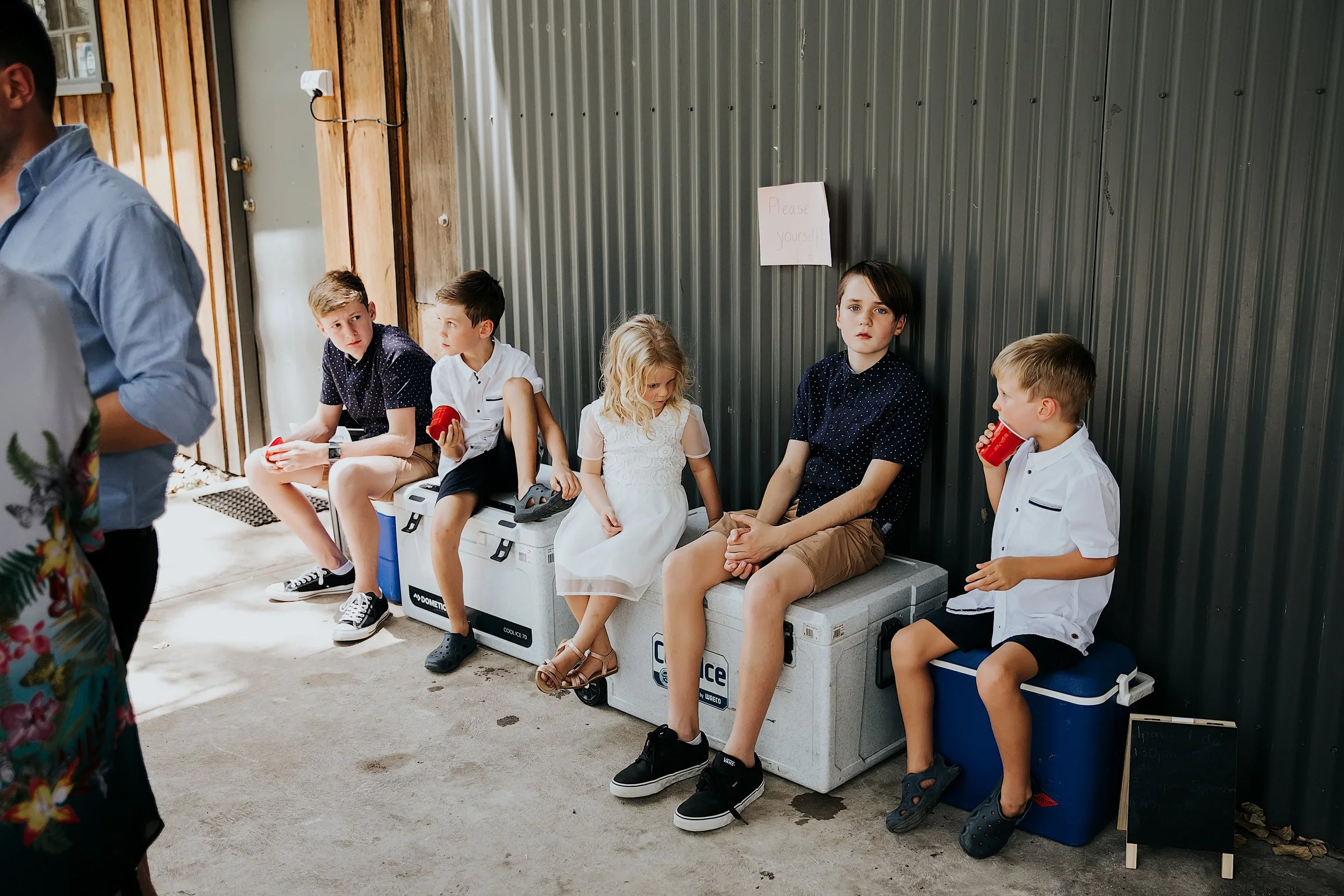 Six children sitting on coolers against a gray metal wall. Four boys and two girls, some with cups, some looking around or at each other. There's a sign that says "Please yourself" on the wall. Part of an adult in floral clothing is visible on the le
