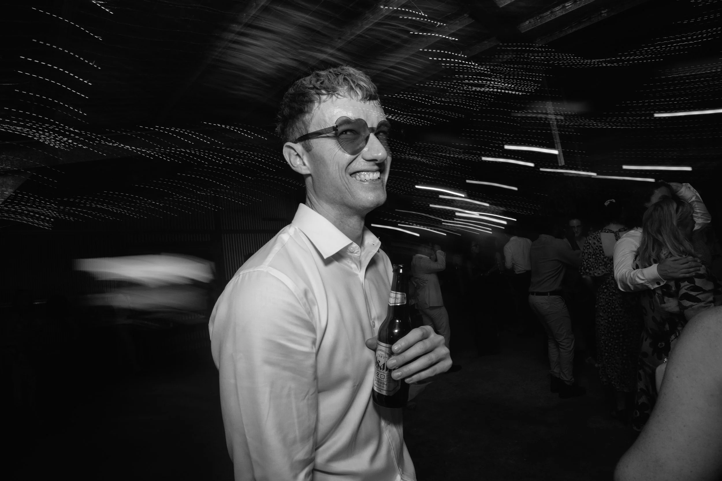 A man with sunglasses holding a beer bottle at a party or social gathering, with blurred lights and other people in the background.