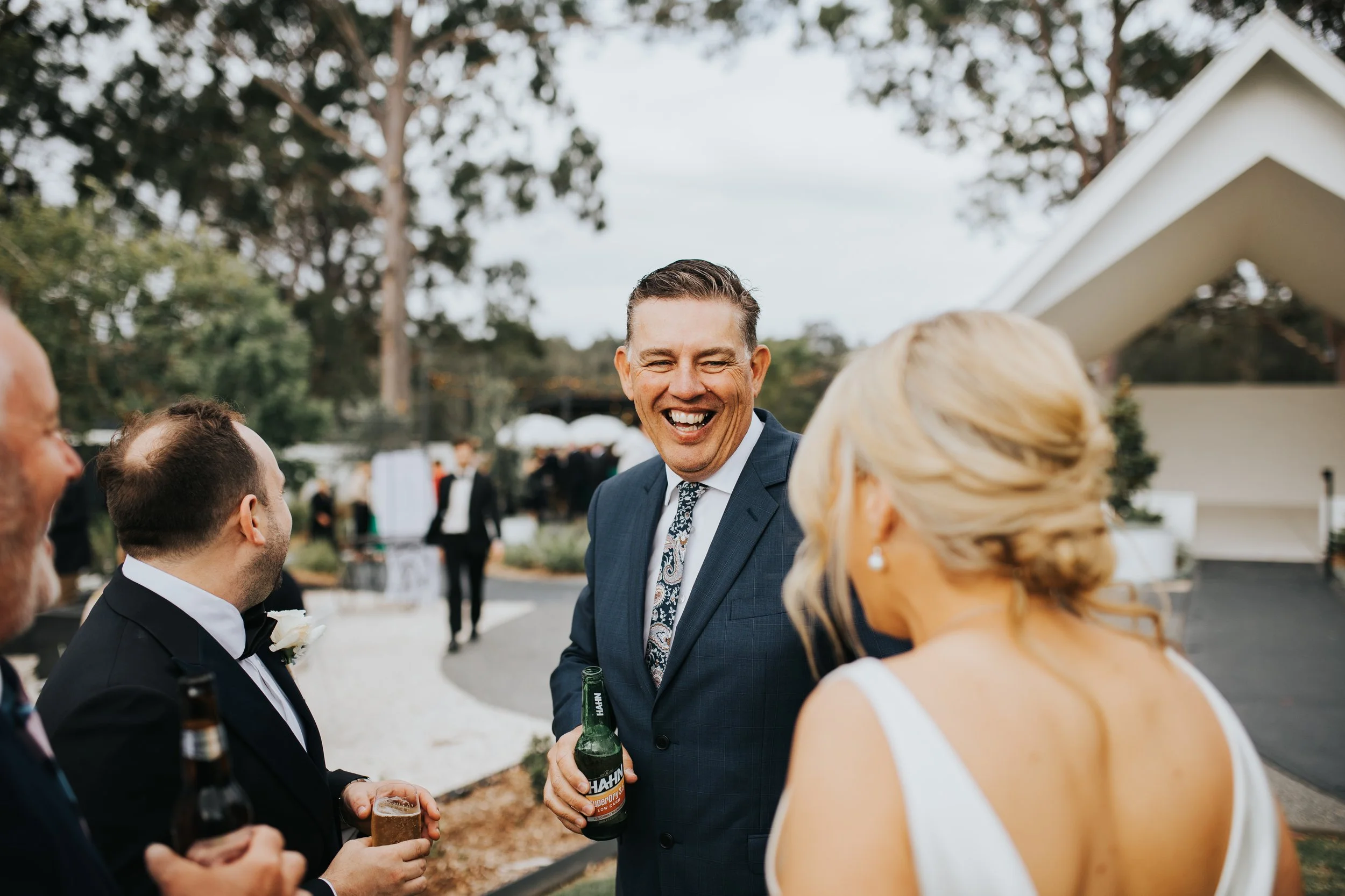 Group of people at an outdoor wedding reception, including a man in a suit with a floral tie holding a beer, and several guests in formal attire, engaging in conversation amid trees and a white building.