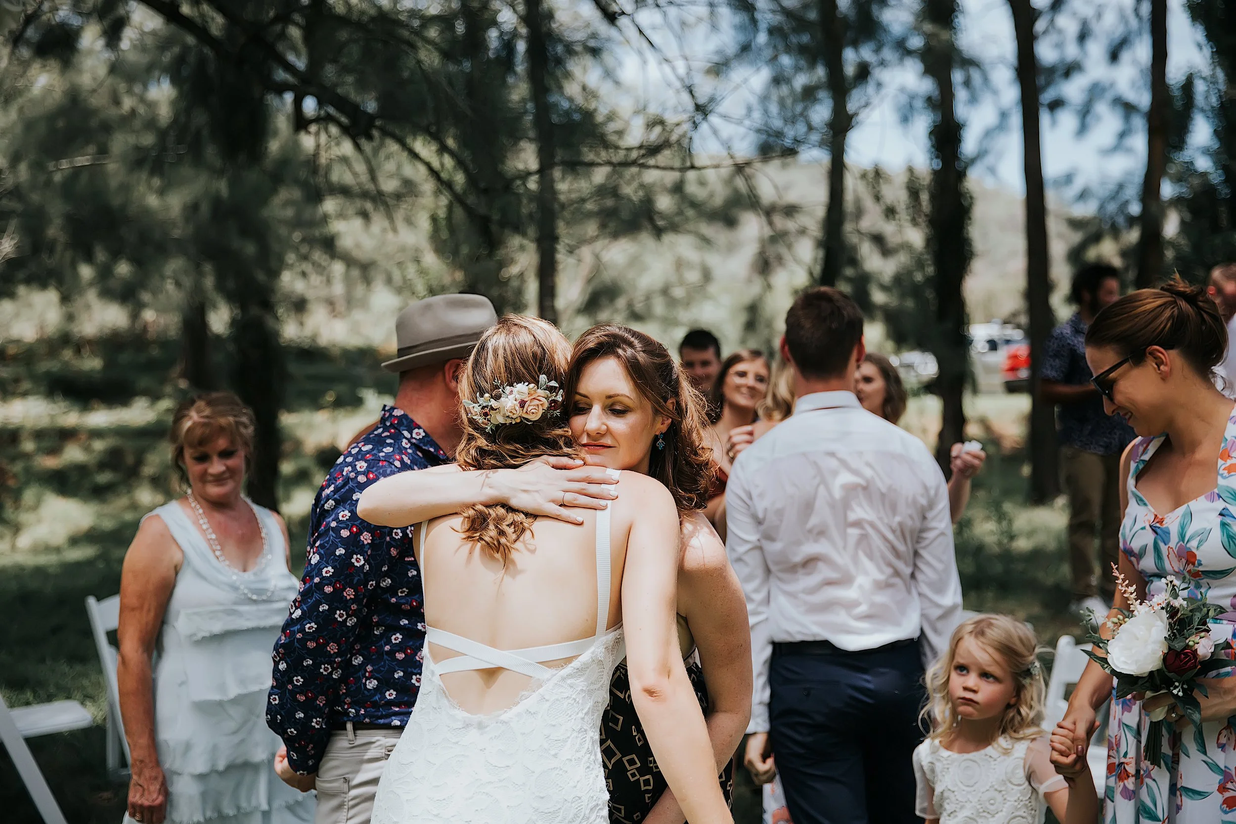 A group of people at an outdoor wedding or celebration in a wooded area, with a woman embracing a man in a floral shirt and a woman in a white dress with a floral hair accessory in the center.