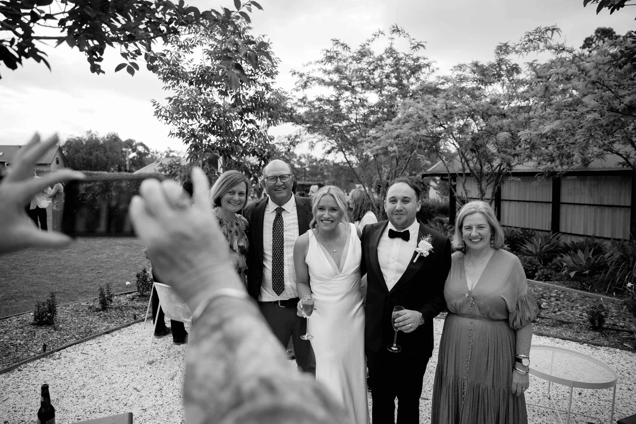 Group of five people, three women and two men, smiling and posing outdoors at a celebration, with trees and a house in the background. One woman is in a white dress holding a glass of champagne. The man next to her is in a tuxedo. The others are dres