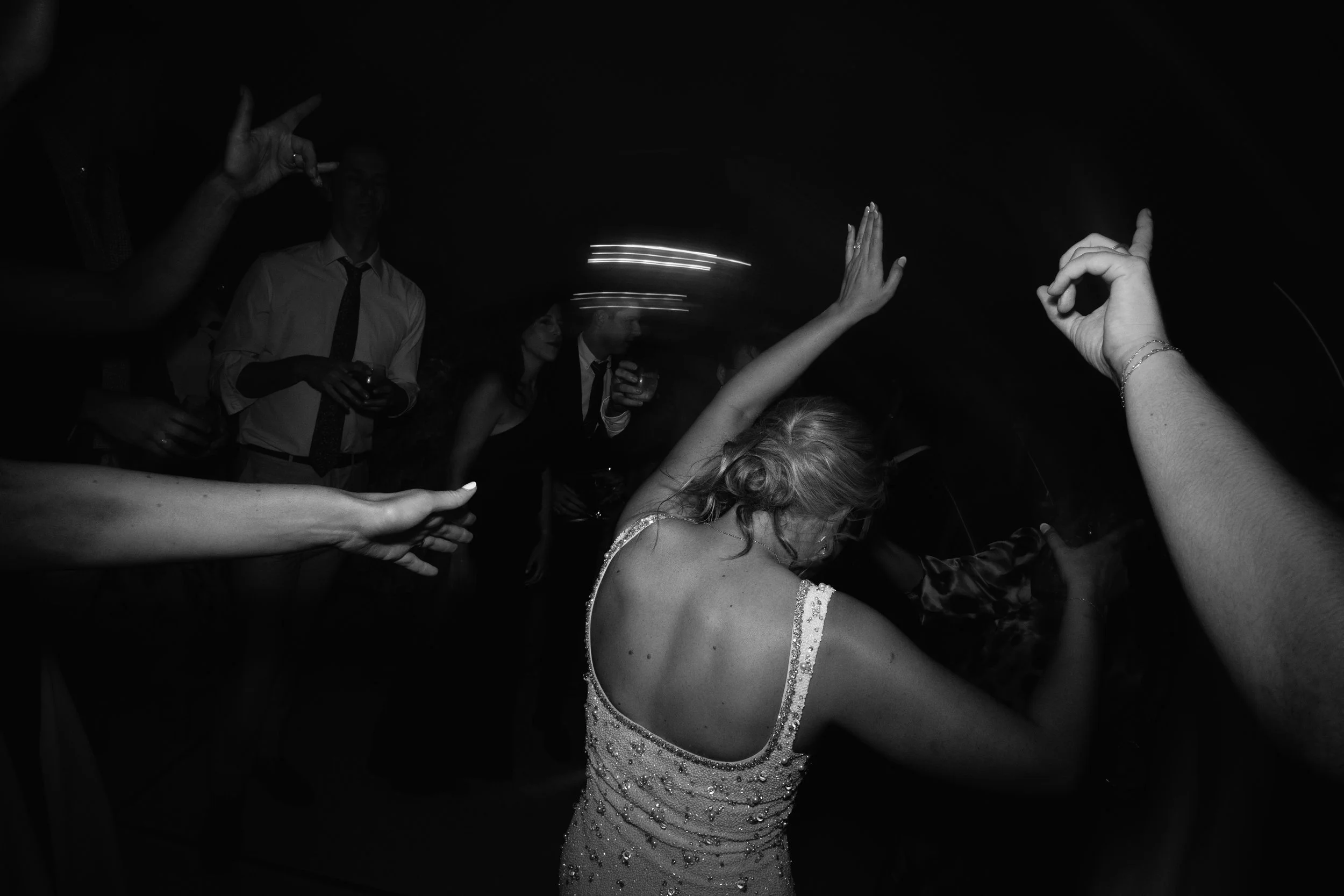 Black and white photo of people dancing at a party, with a focus on a woman in a sparkly dress and adigitized light streaks in the background.
