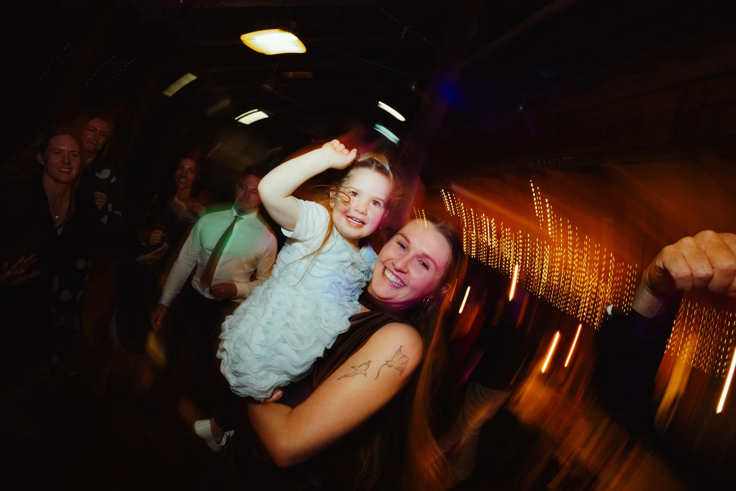 A woman and a young girl smiling and dancing at a party with colorful lights and other guests in the background.