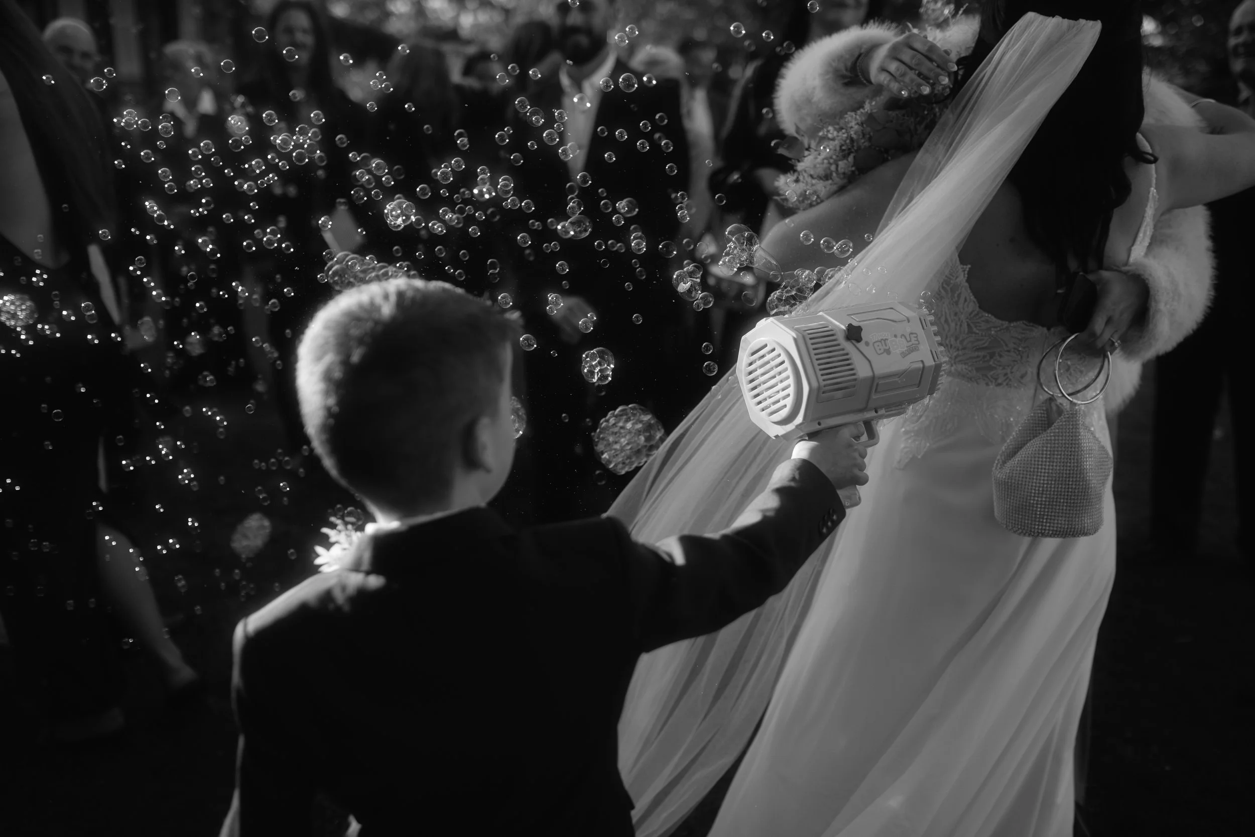 A bride and a groom embrace during their wedding celebration, with bubbles floating around them and guests in the background.