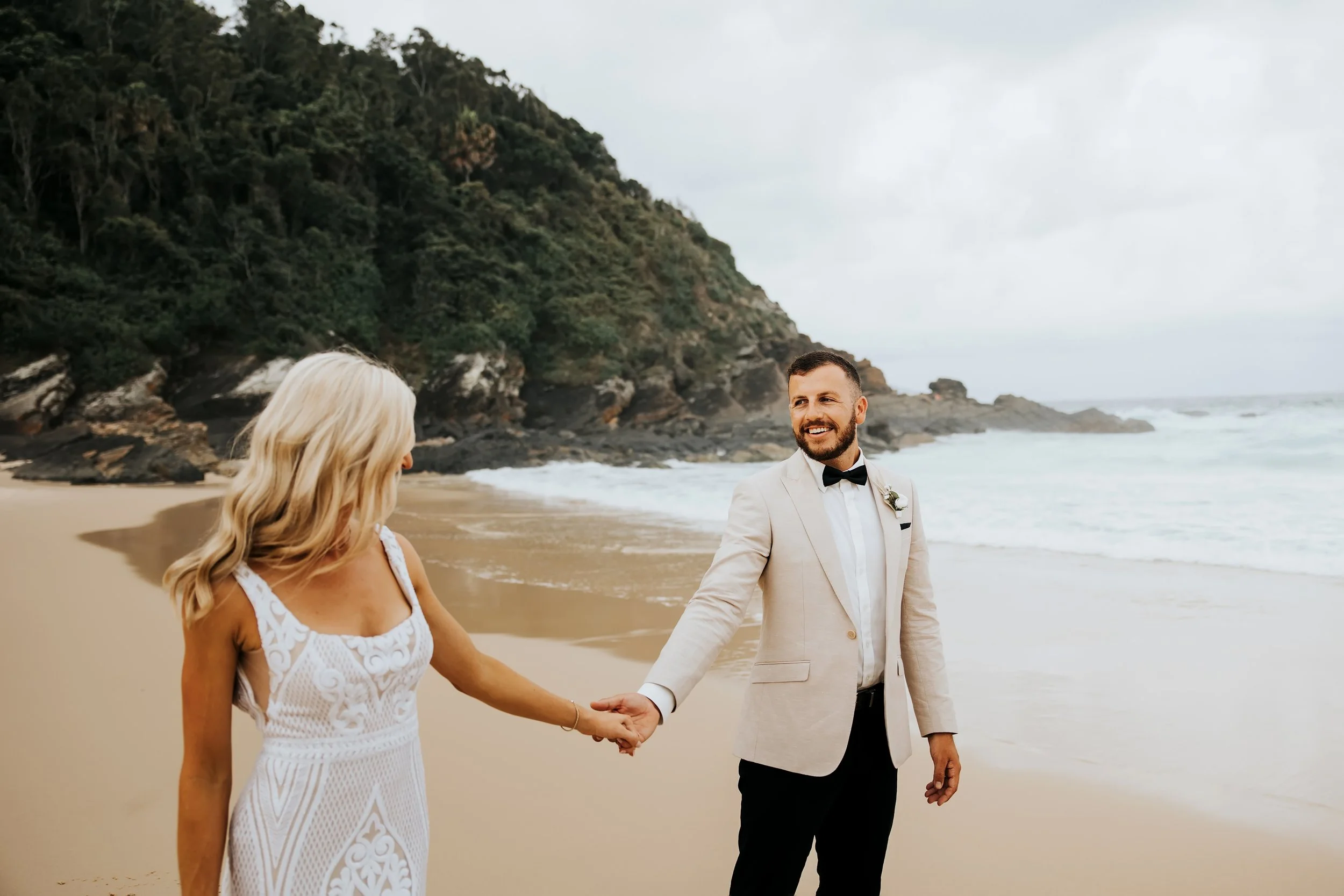 A couple gets married on a beach with cliffs in the background. The groom is in a white suit with a black bow tie, and the bride, seen from behind, is in a white lace dress. They are holding hands and smiling at each other near the shoreline.