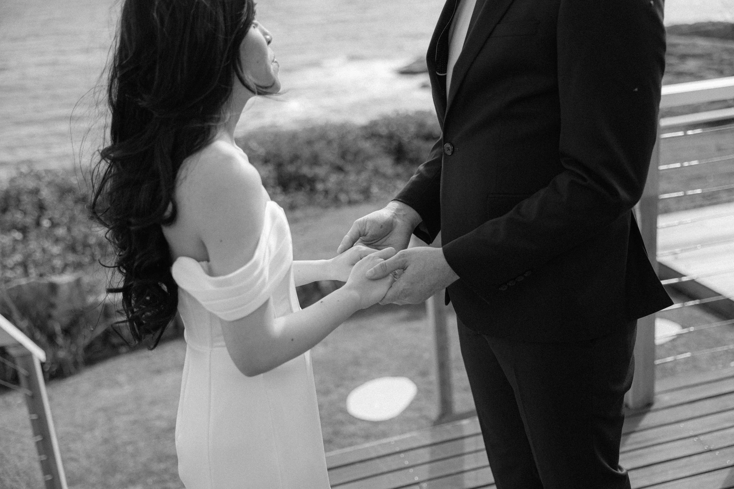 A couple holding hands during a wedding ceremony outdoors by the water, with the bride wearing a strapless dress and long dark hair, and the groom in a suit.