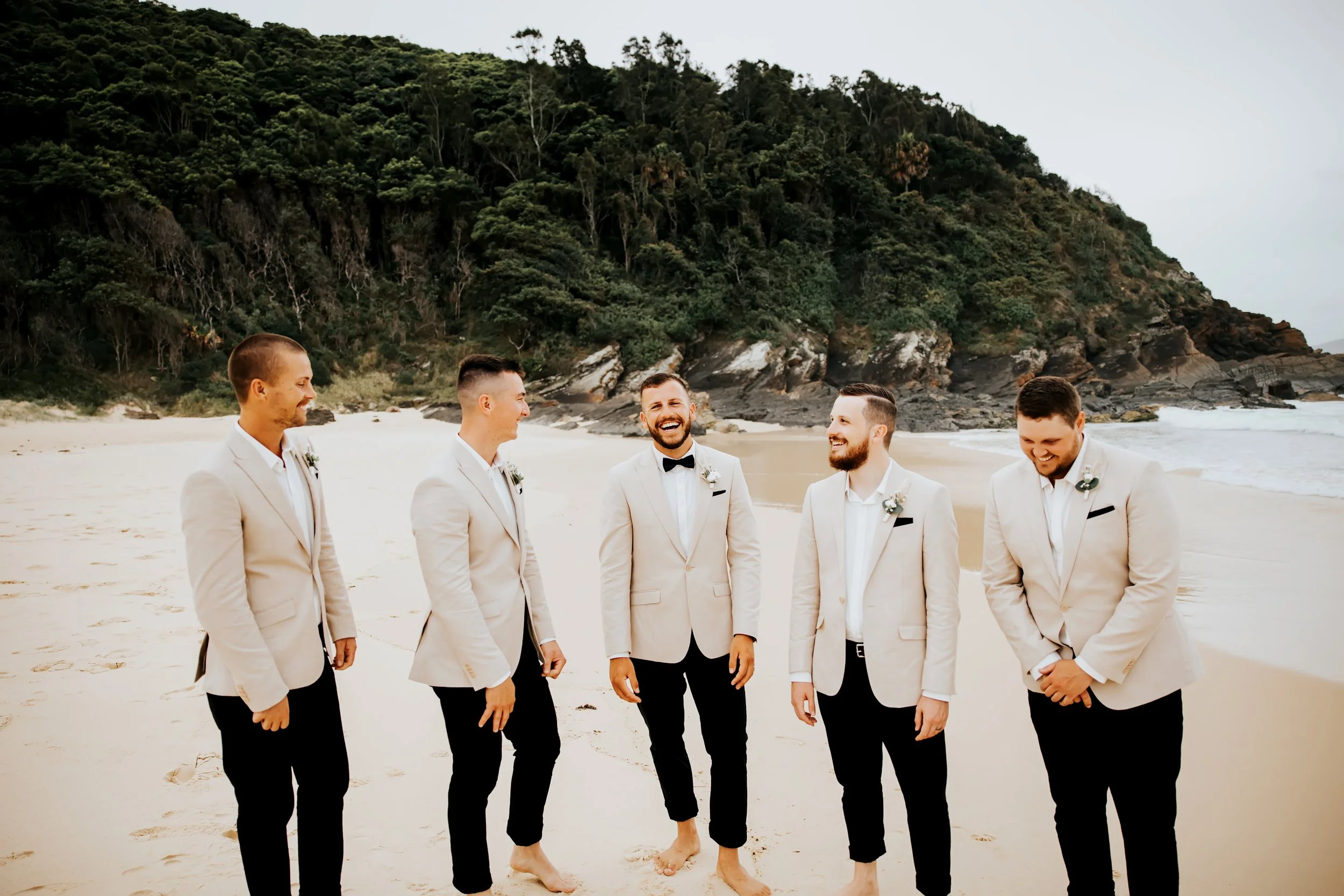 Six men in beige suits and black bow ties standing on a sandy beach with a large green hill and rocks in the background, smiling and enjoying themselves.