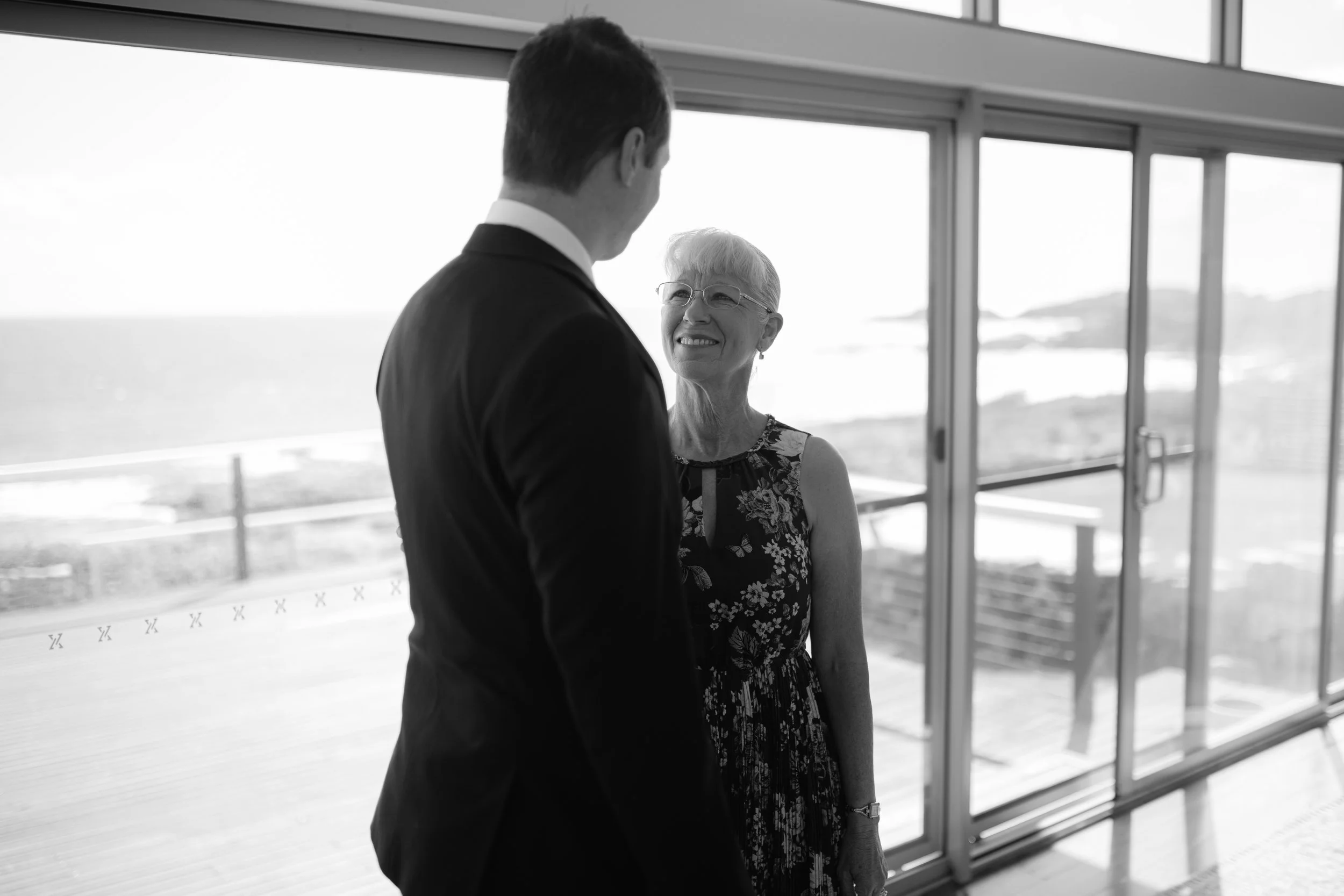 A man in a suit talking to a smiling woman in a floral dress inside a building with large windows overlooking a beach.