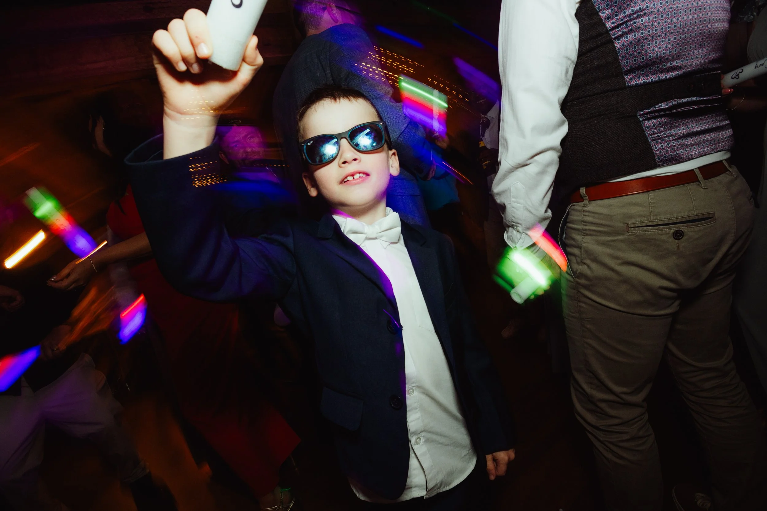 A young boy wearing sunglasses and a tuxedo, taking a selfie at a party with colorful lights and other people dancing around him.