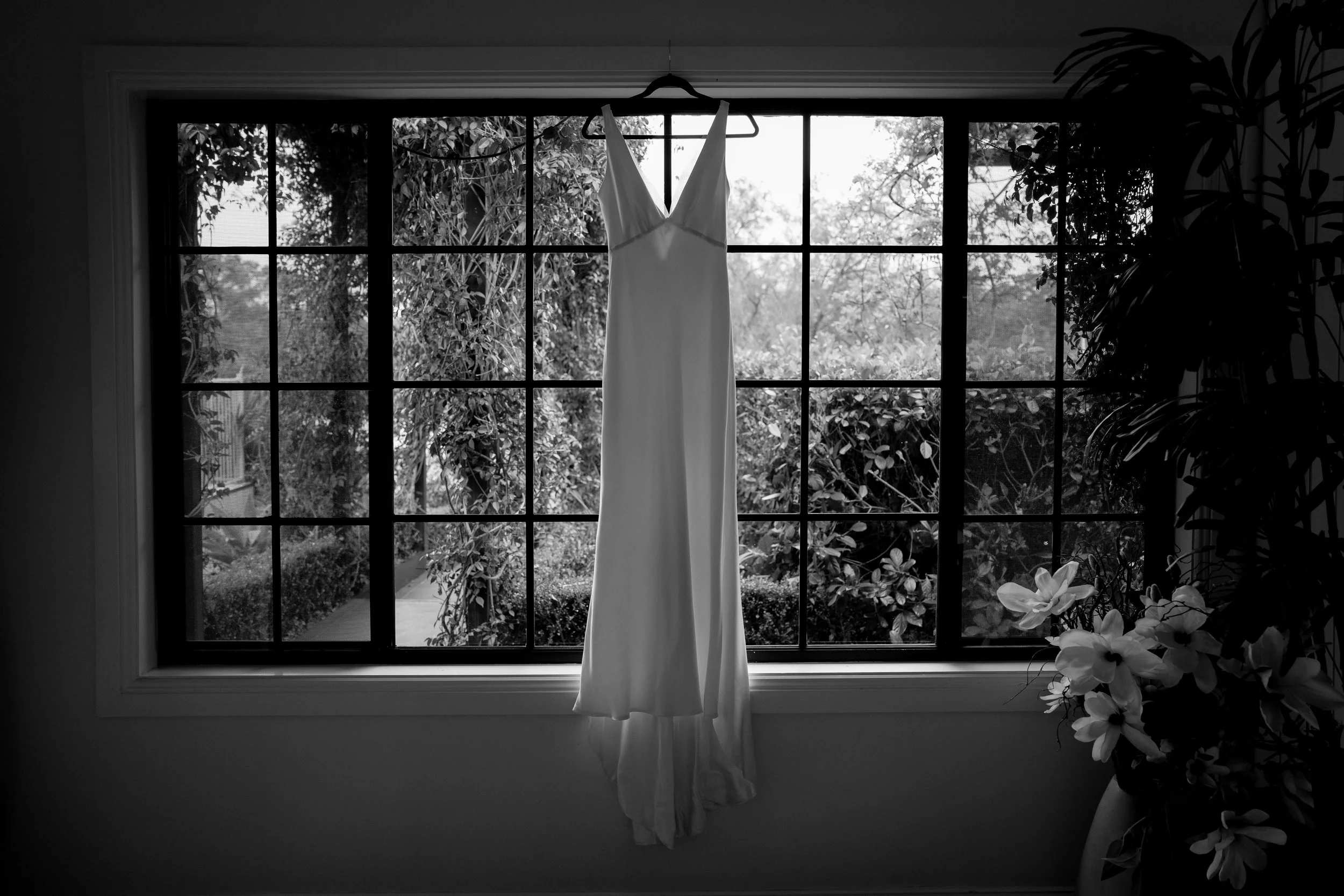 A white wedding dress hanging on a black hanger in front of a large grid window. Outside, there is lush greenery and trees. To the right, a pot of white flowers is visible.
