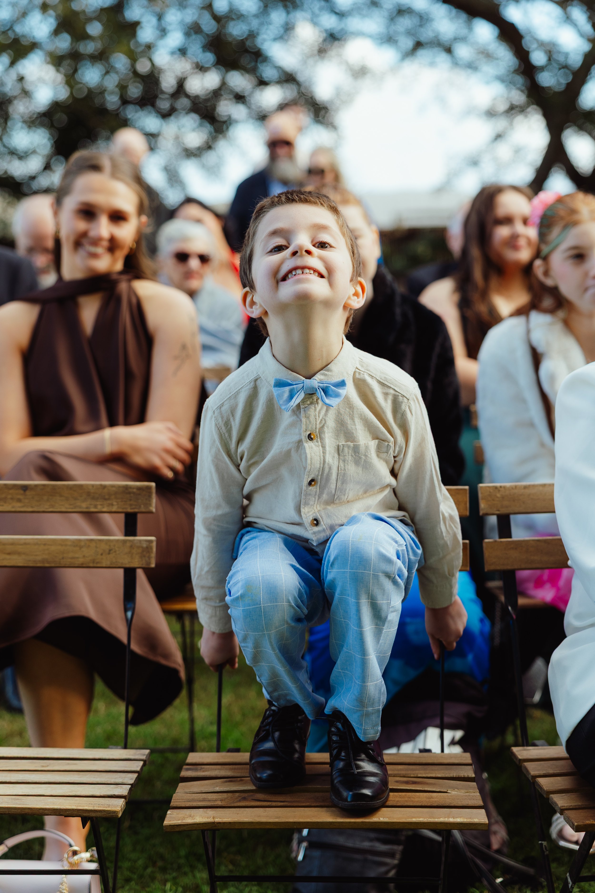 A young boy with a big smile, wearing a beige shirt, blue bow tie, and blue plaid pants, sitting on a wooden chair outdoors during daytime, surrounded by adults and children.