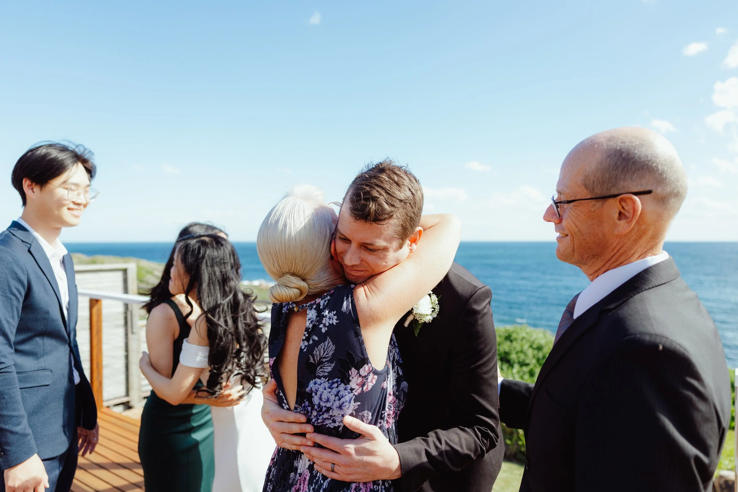 People gathered outdoors by the ocean, hugging and smiling during a sunny day with a clear blue sky.