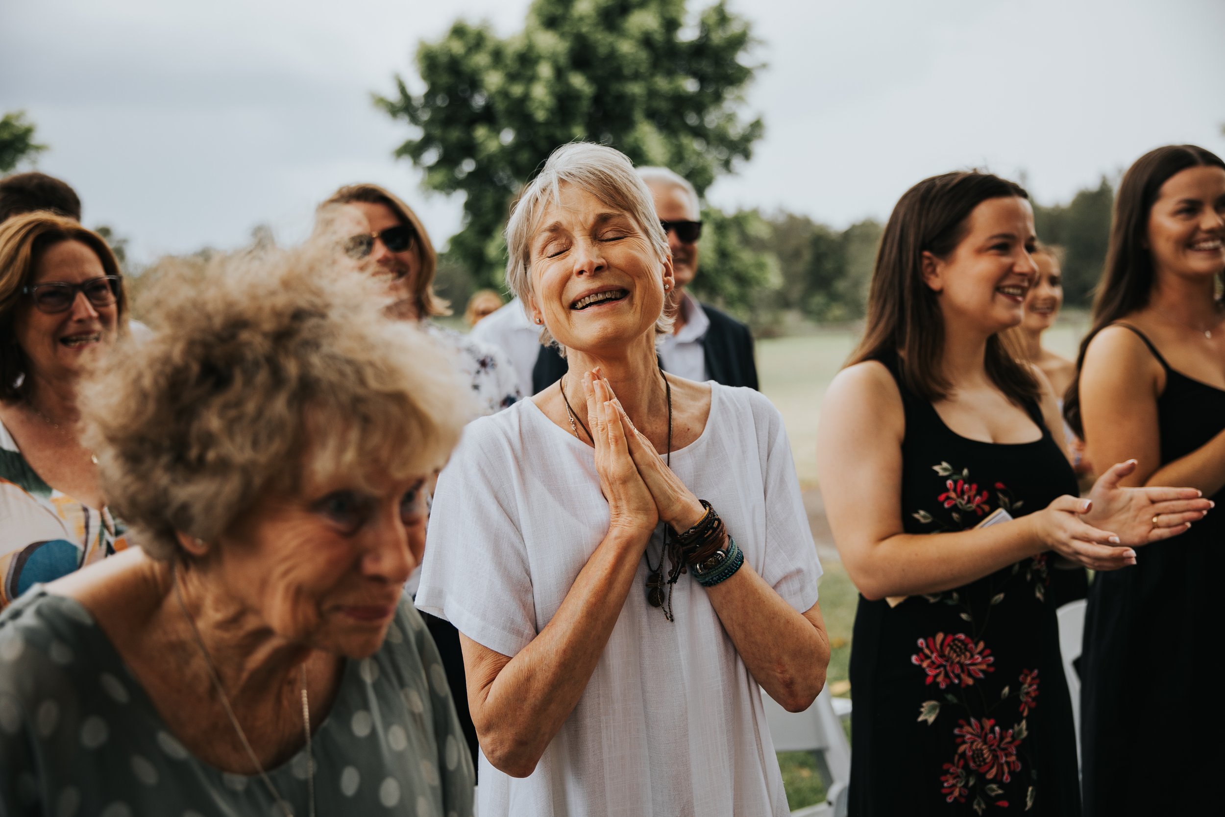 A group of people outdoors, with an elderly woman at the center with eyes closed, smiling, hands pressed together in prayer or gratitude, surrounded by smiling others.