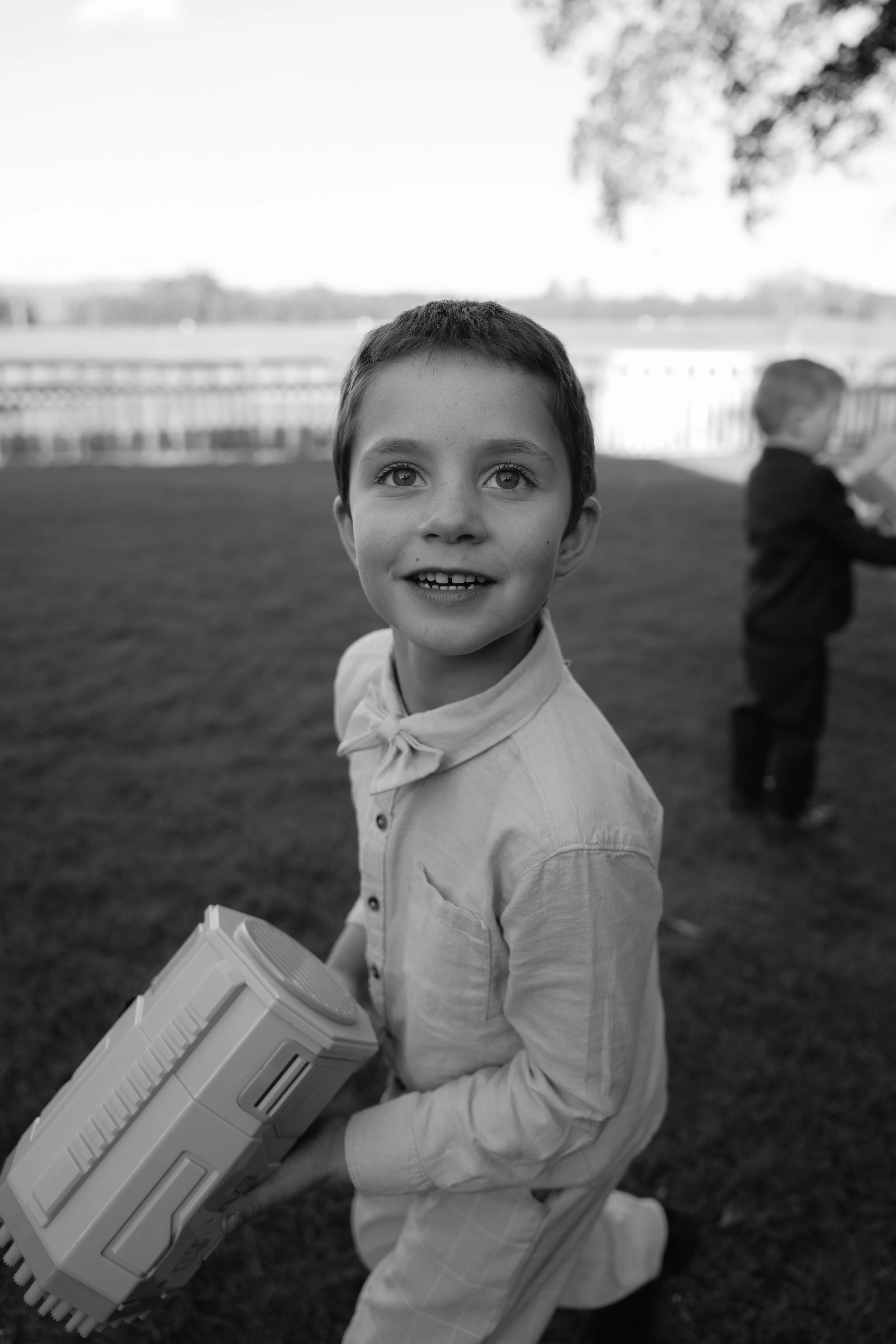 A young boy with short hair and a striped shirt holding a toy gun outdoors on grass, with another child in the background holding an object, blurred with trees and open sky behind them, in black and white.