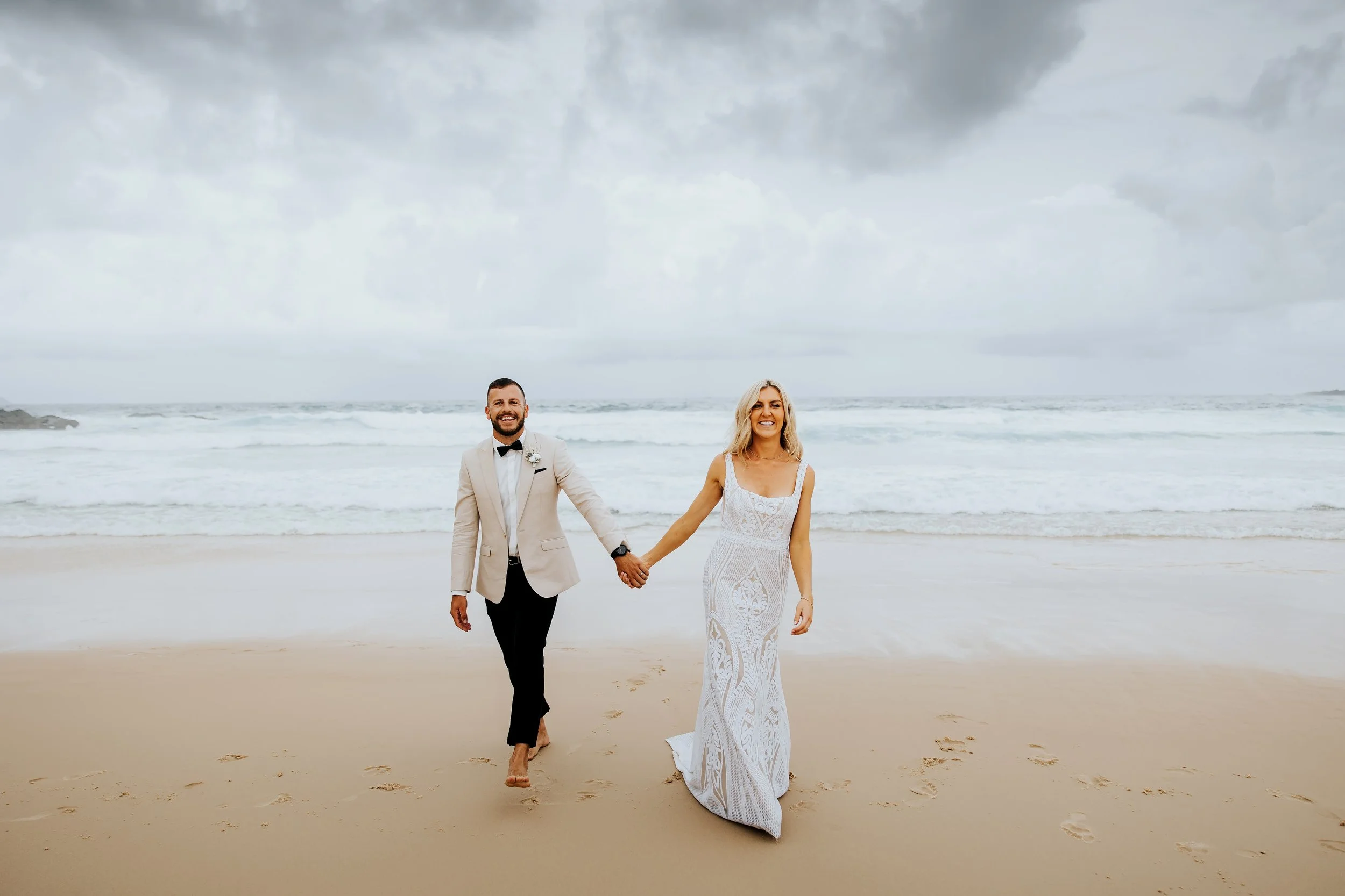 A smiling couple, dressed in wedding attire, holding hands and walking barefoot along the beach with waves and cloudy sky in the background.