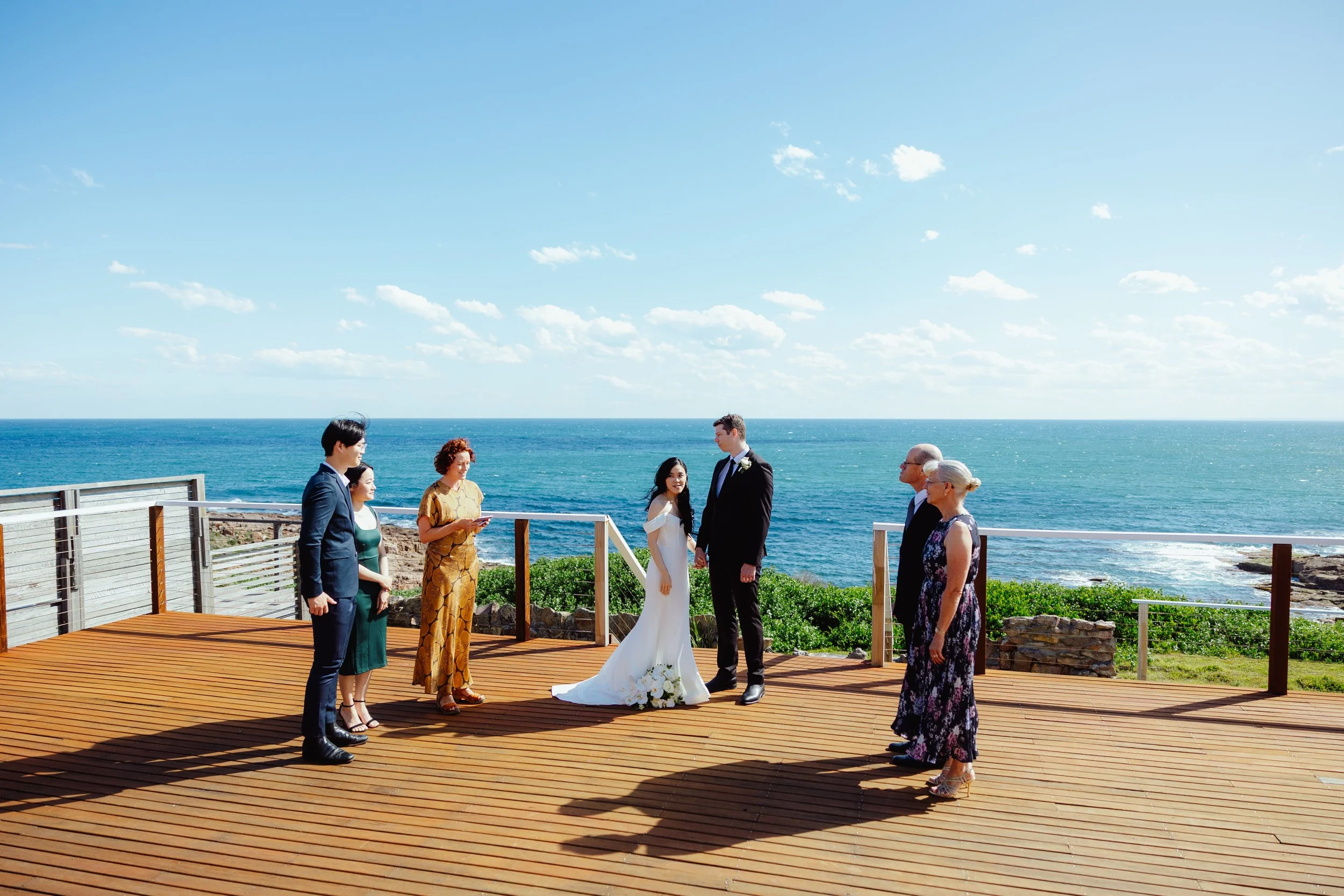 A wedding ceremony taking place on a wooden deck by the ocean with a clear blue sky. The bride and groom are standing in the center facing each other, surrounded by family and friends.