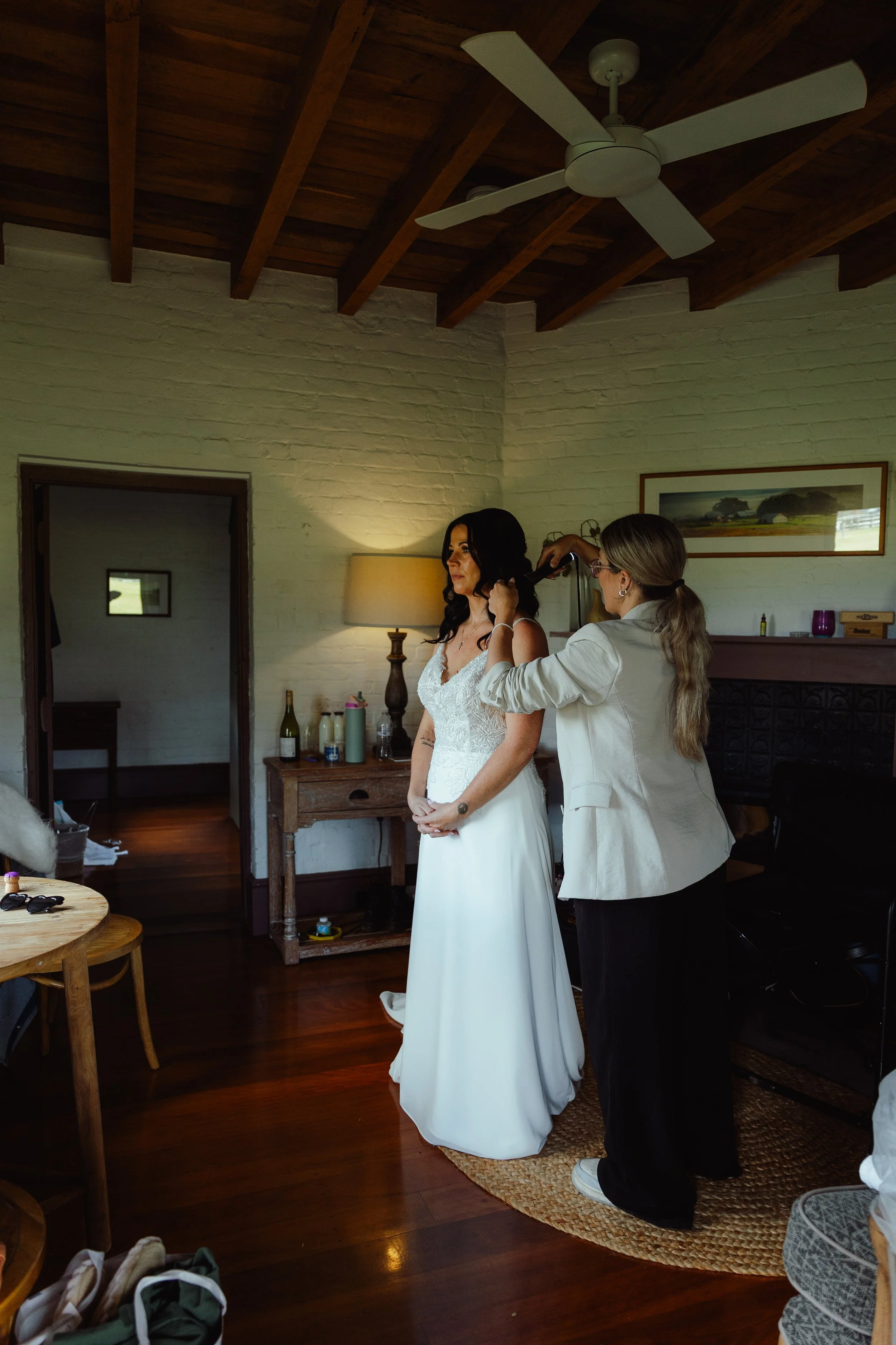 A woman in a white wedding dress is being hair styled by another woman in a white blazer, in a cozy room with wooden exposed beams and white brick walls.