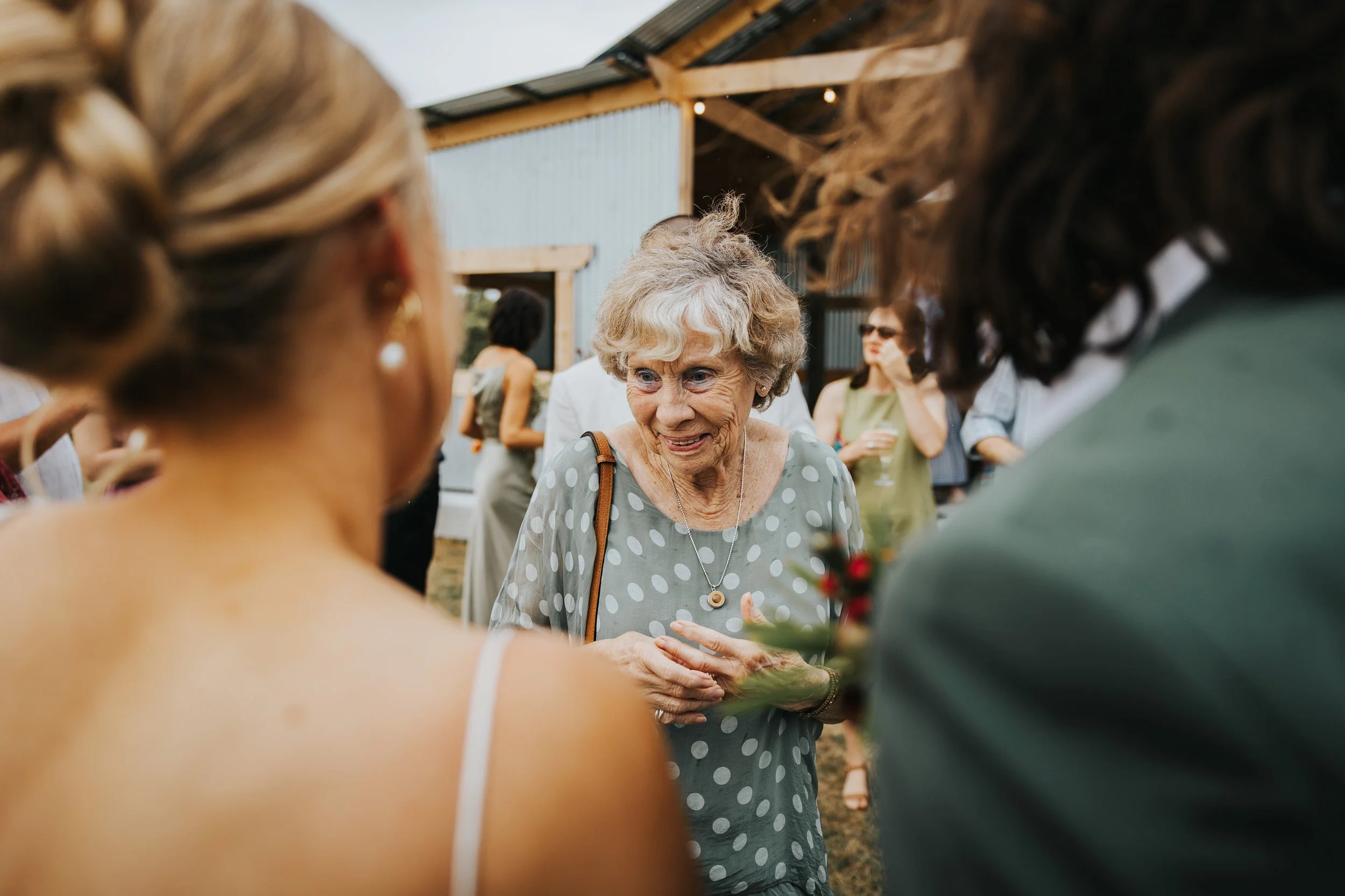 A group of people socializing outdoors, centering on an elderly woman with short gray hair, wearing a gray dress with white polka dots, smiling and talking. The background shows more people, a barn-like structure, and a partly cloudy sky.