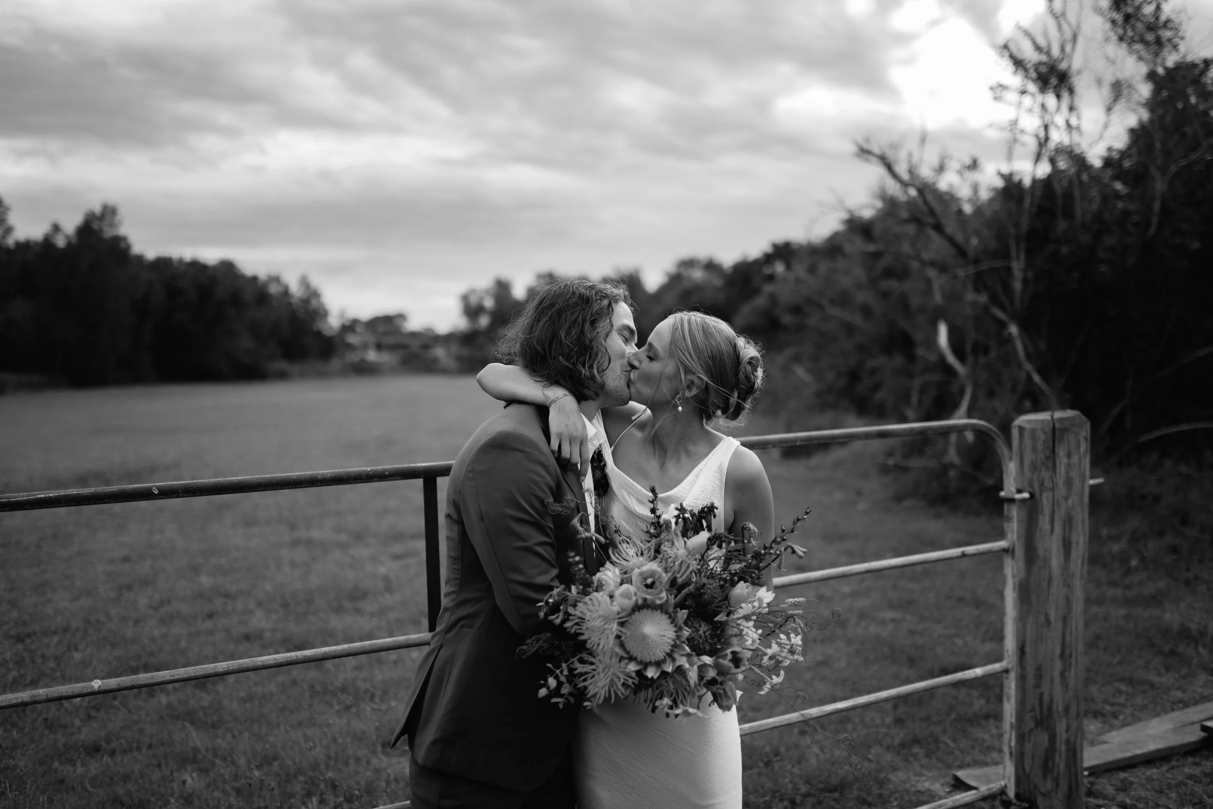 A black and white photo of a couple sharing a kiss outdoors near a fenced area with trees and cloudy sky in the background. The woman is holding a bouquet of flowers.